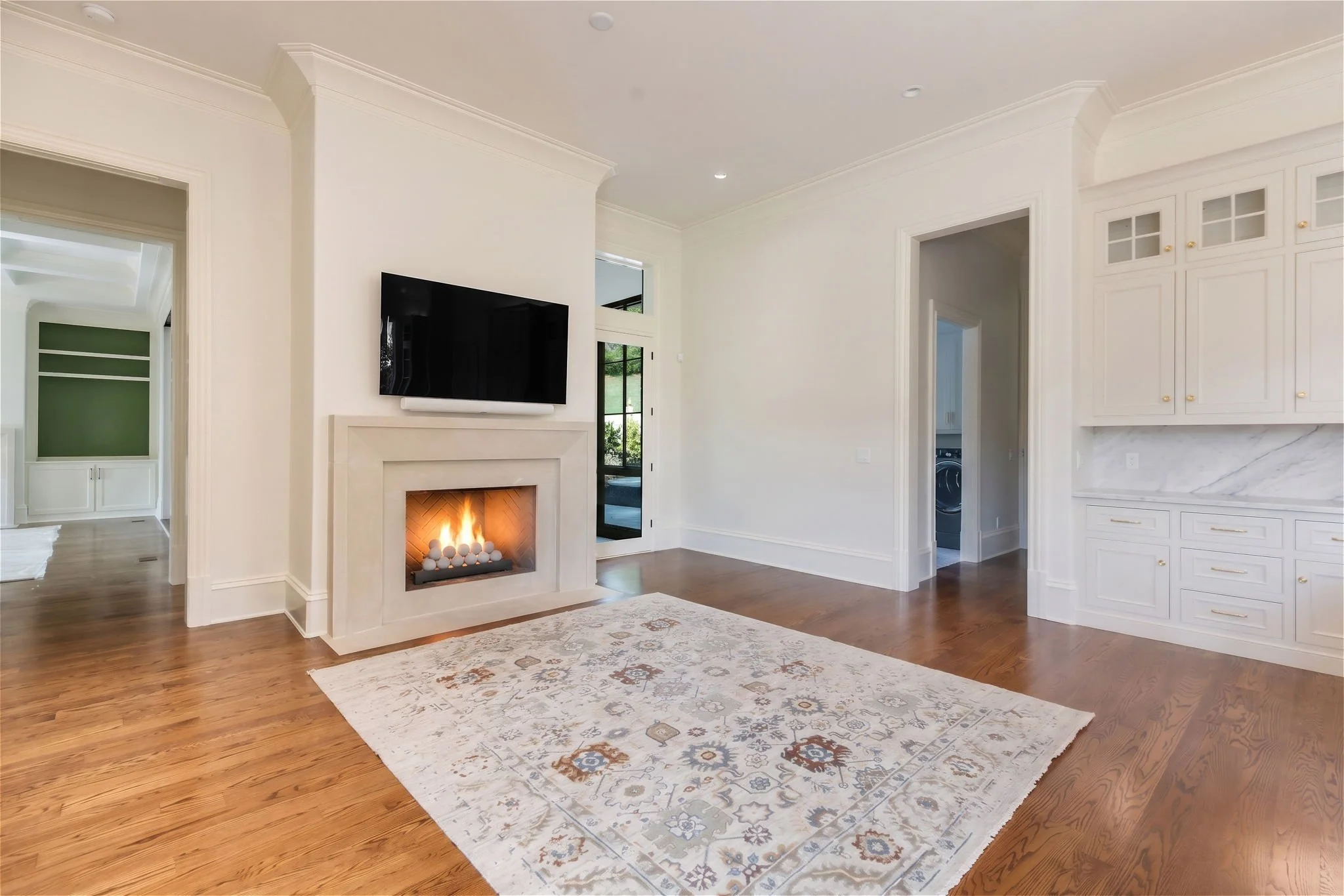 Living room with hardwood floors, a white fireplace with a black screen, a wall-mounted flat-screen TV above the fireplace, a patterned area rug, white cabinetry on the right, and a doorway leading to a laundry room.