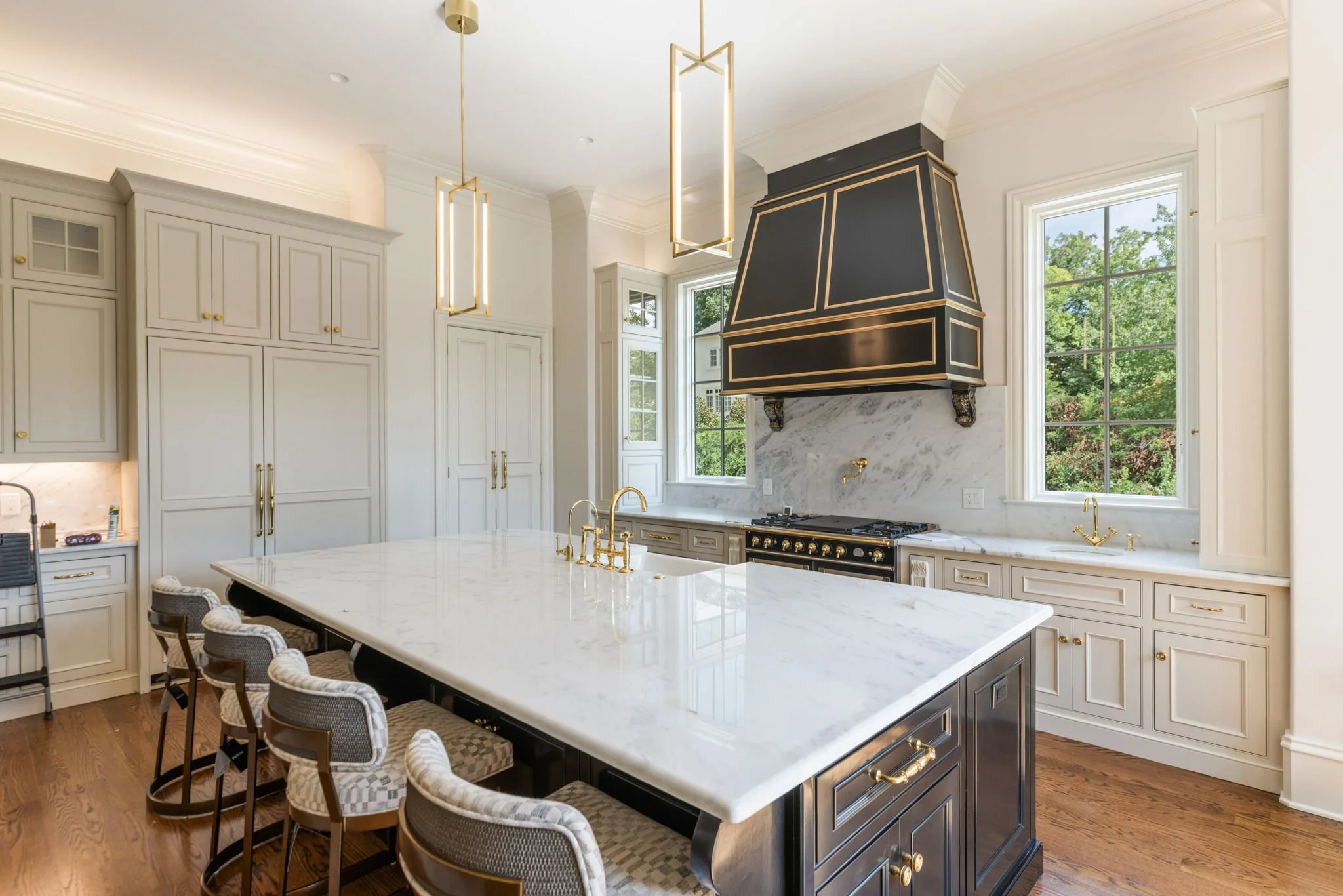 A modern kitchen with white cabinets, black appliances, and gold accents. It features a large marble island with gold fixtures and stylish chairs, gold pendant lights, large windows with a view of green trees, and a black range hood with gold trim ab