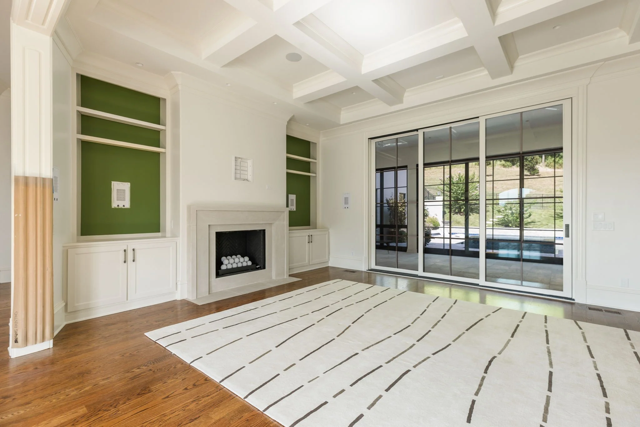 Living room with high coffered ceiling, green accent wall above a fireplace, large sliding glass door leading outside, and hardwood floors covered partially by a beige area rug with black lines.