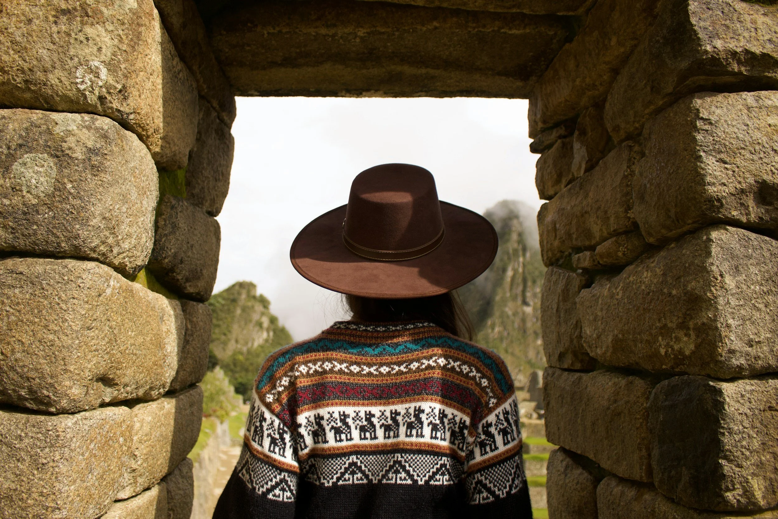 Person with long hair wearing a colorful patterned sweater and a wide-brimmed brown hat, viewed from behind, standing in a stone border opening with a landscape of greenery and rocky formations in the distance.