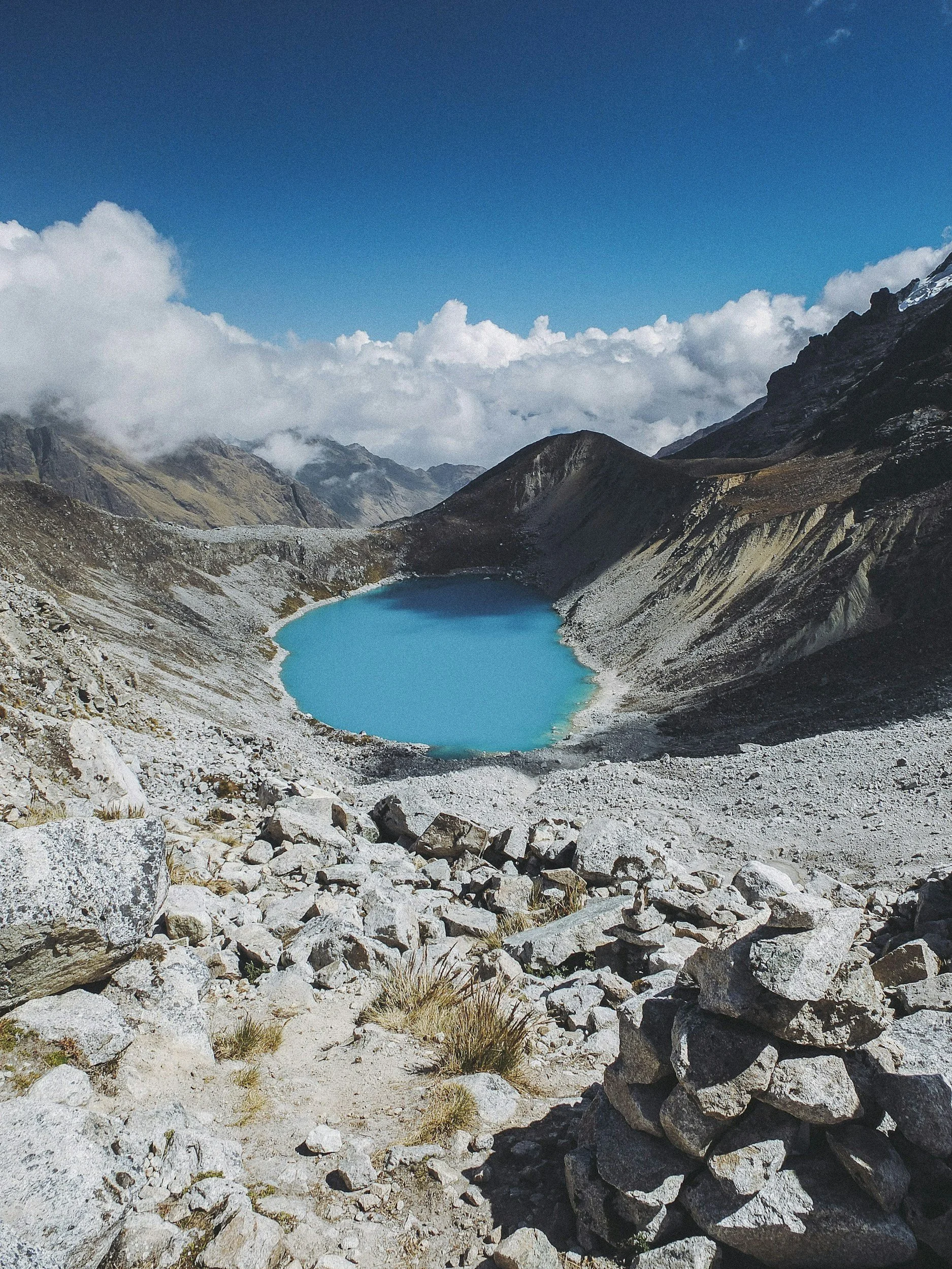 Scenic mountain landscape featuring a small turquoise lake surrounded by rocky terrain and high mountain peaks under a partly cloudy sky.