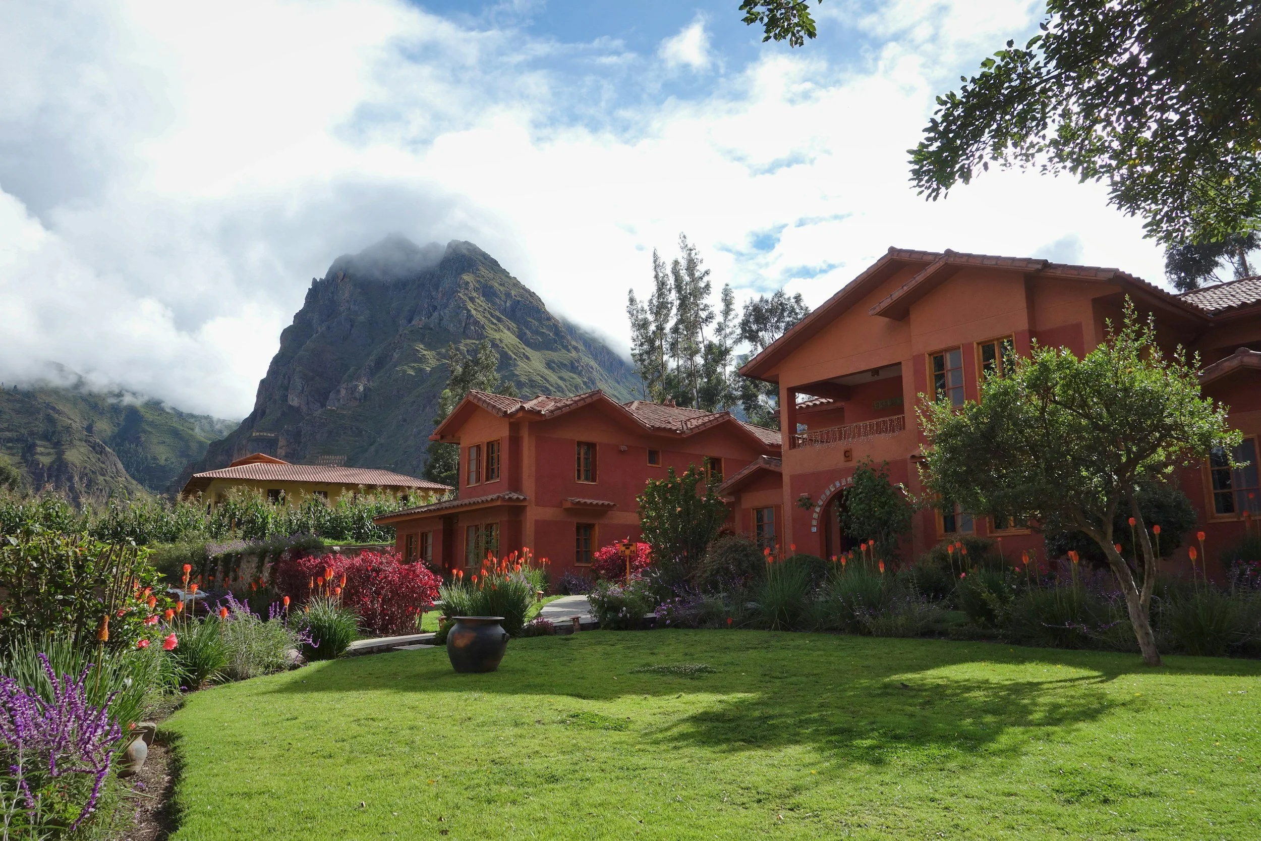 A house with red walls and a red tiled roof surrounded by a lush green lawn and colorful flowers, with tall mountains and cloudy sky in the background.