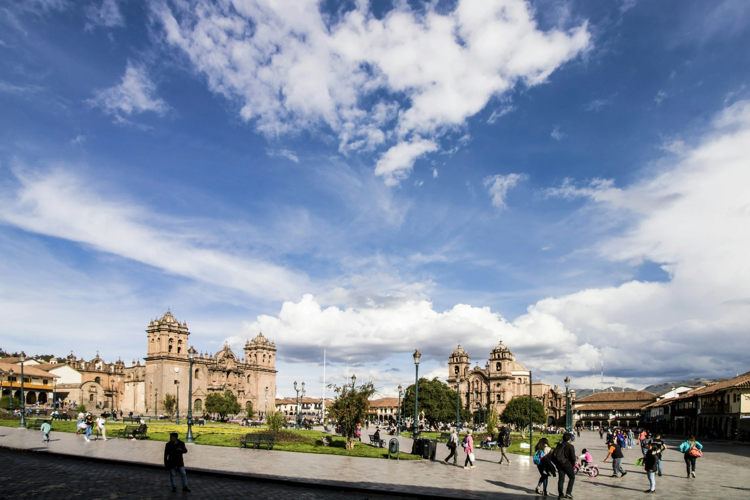 Plaza with people walking and sitting, historic pink stone churches, green lawns, lampposts, under a partly cloudy blue sky.