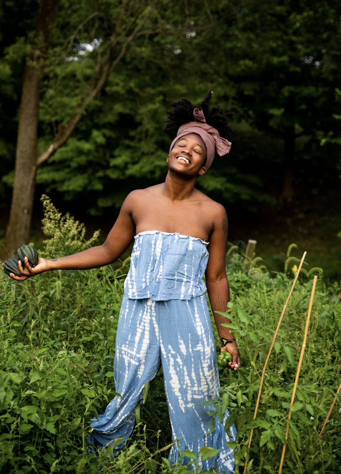 A woman in a tie-dye blue and white strapless jumpsuit and a pink head wrap is smiling happily in a garden, holding dark green zucchini in her hand.