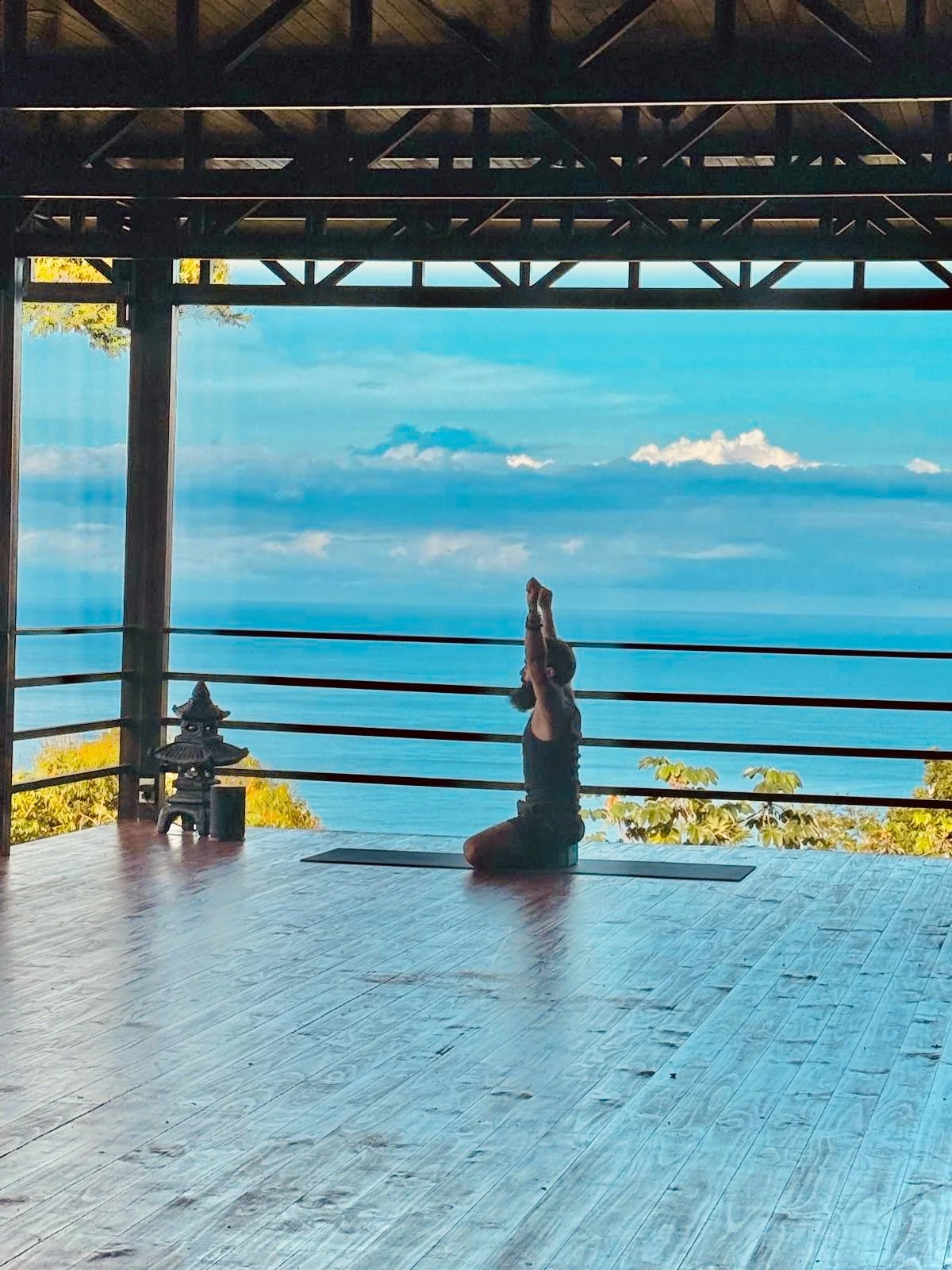 A person practicing yoga in a kneeling pose with arms raised in front of an open-air wooden structure overlooking the ocean. The scene includes a yoga mat, a small lantern, and a background of blue water and sky with clouds.