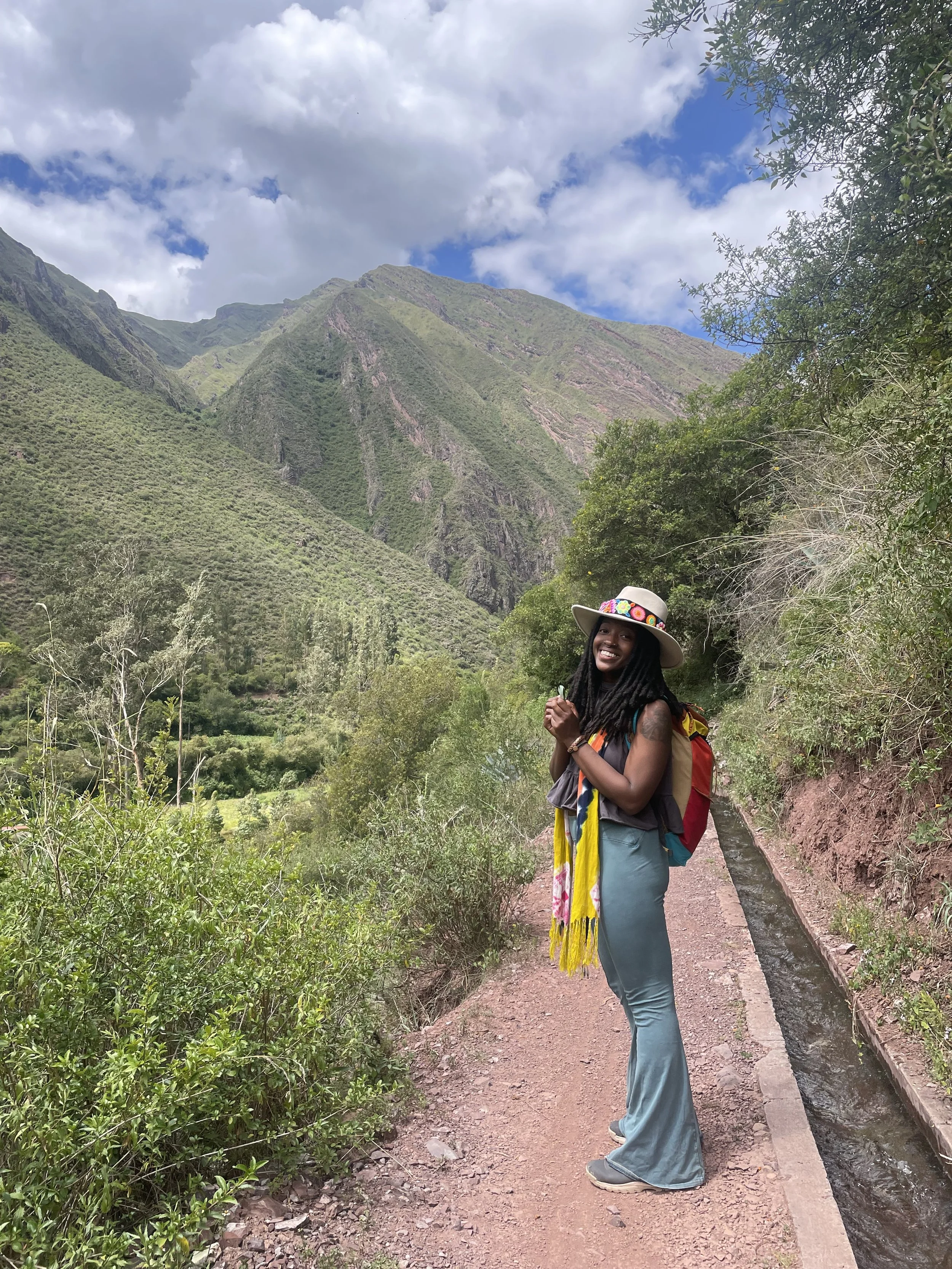 A woman hiking along a mountain trail with lush greenery and tall mountains in the background, wearing a hat, colorful scarf, and carrying a backpack.