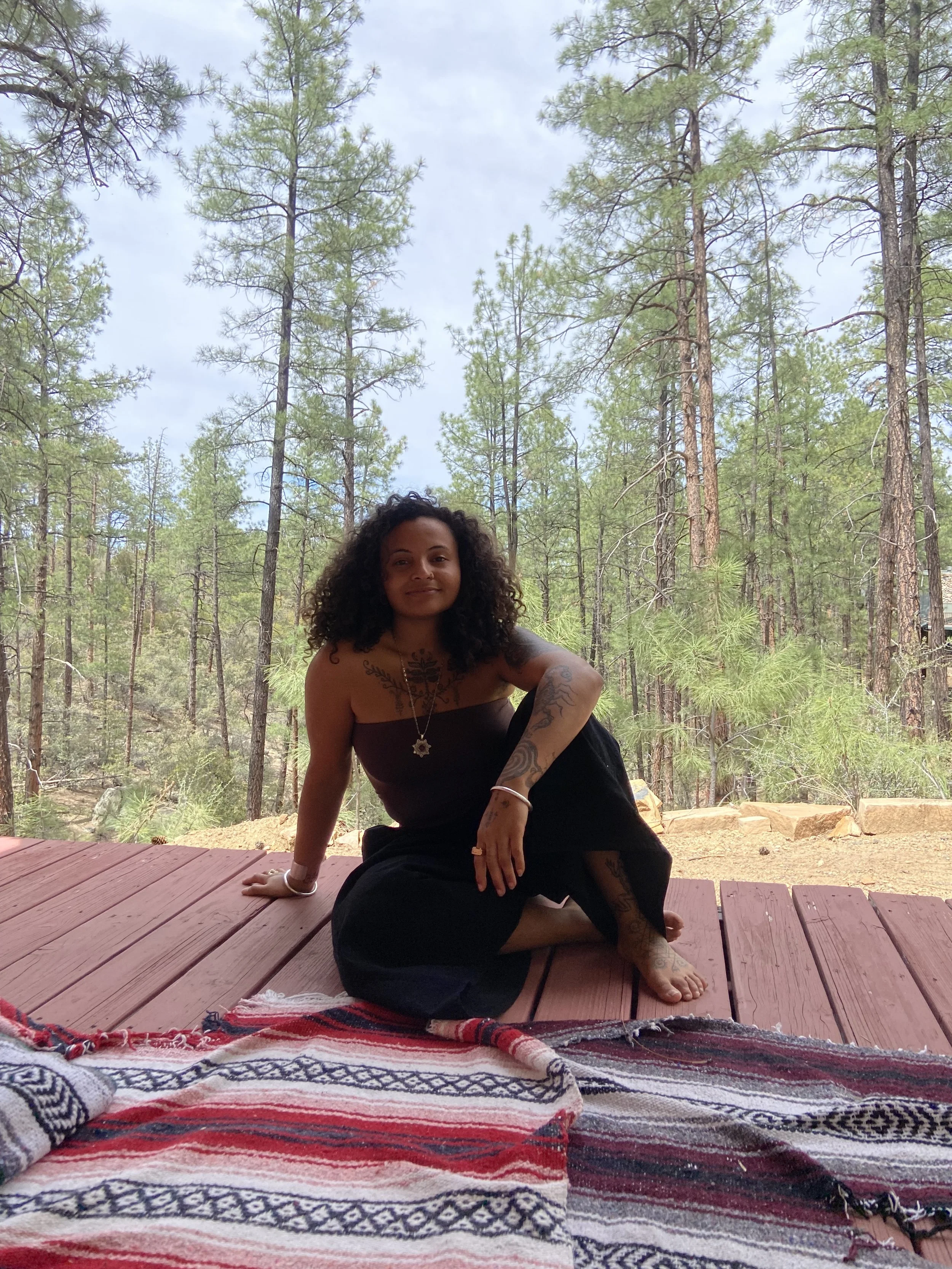 A woman with dark curly hair sitting cross-legged on a wooden porch with a forest of pine trees in the background. She is wearing a strapless black top and black pants, with tattoos on her arms and chest, along with jewelry including a necklace and bracelet. There is a colorful striped blanket on the porch in the foreground.