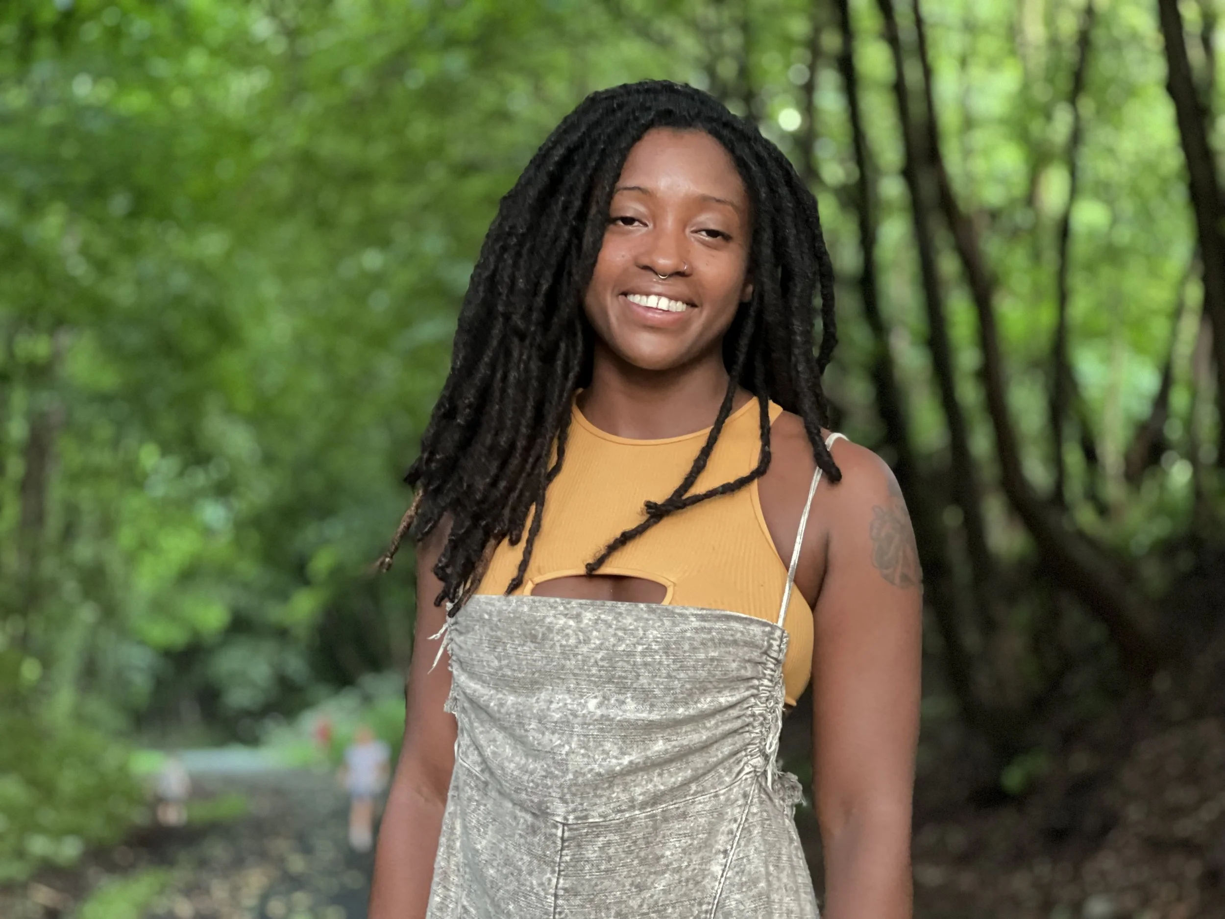 A smiling woman with long dreadlocks standing outdoors on a lush green forest trail.