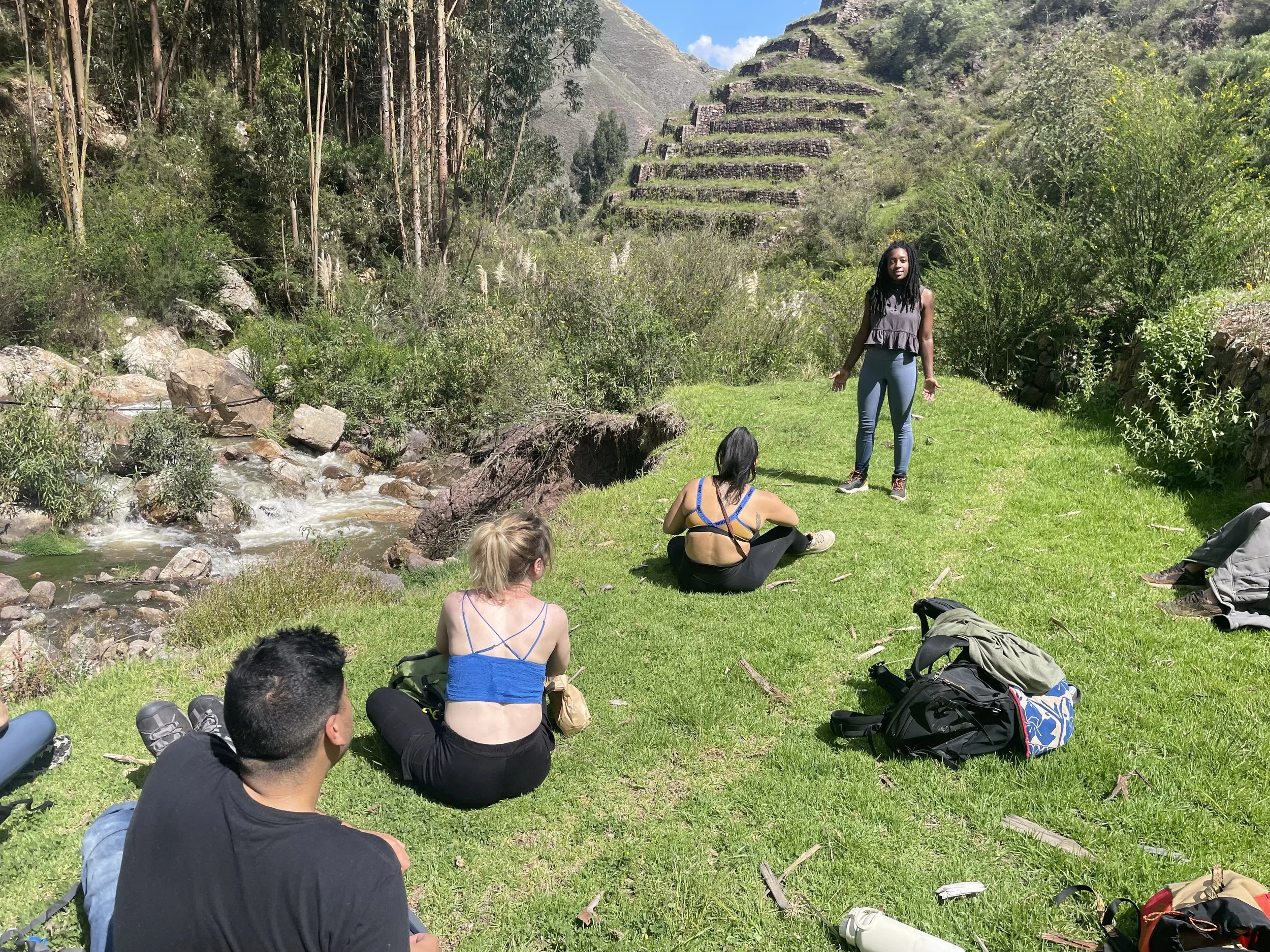 Group of people sitting and standing on grassy area beside a flowing creek, with lush green trees and a stepped hillside in the background on a sunny day.