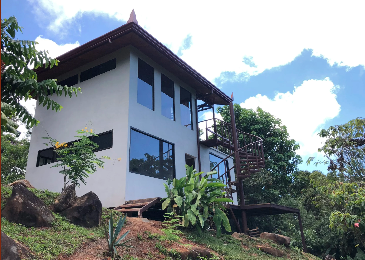 Modern two-story house with white walls and large windows, surrounded by greenery and rocks, under a partly cloudy sky.