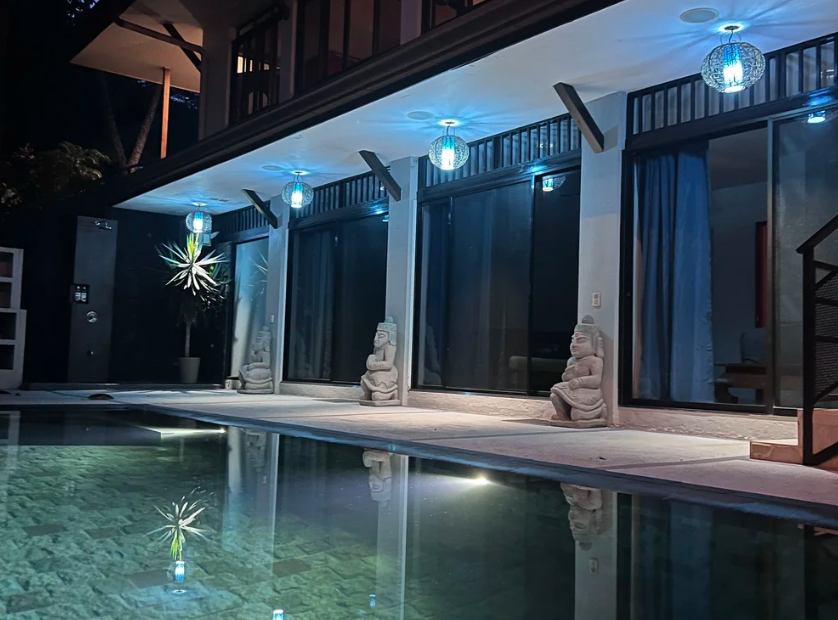 Nighttime view of a modern house with a swimming pool, decorated with blue hanging lanterns, stone statues on the patio, and large glass doors with curtains.