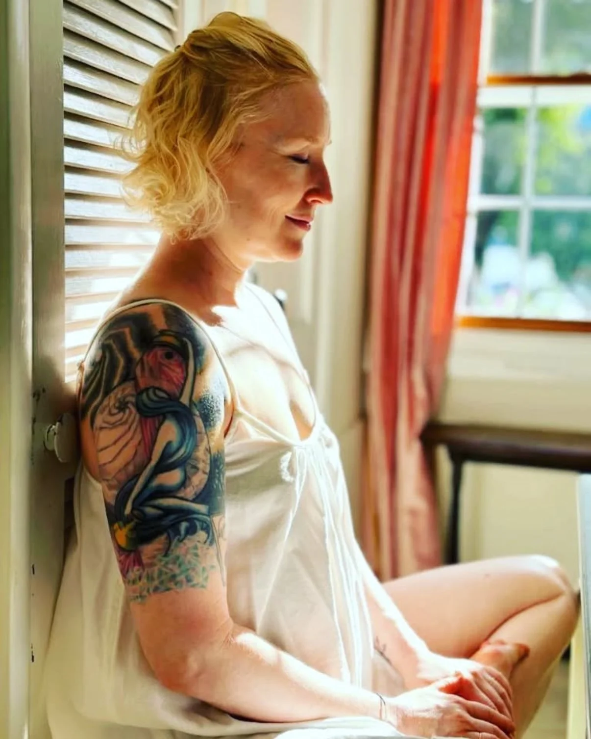 A woman with blonde hair and colorful tattoos on her arm, sitting with her legs crossed and eyes closed, leaning against a wall near a window with curtains.