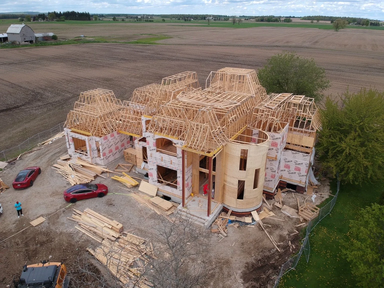 A large house under construction with a wooden framework and roof trusses, surrounded by construction materials, vehicles, and a fenced yard, in a rural area with open fields and trees.
