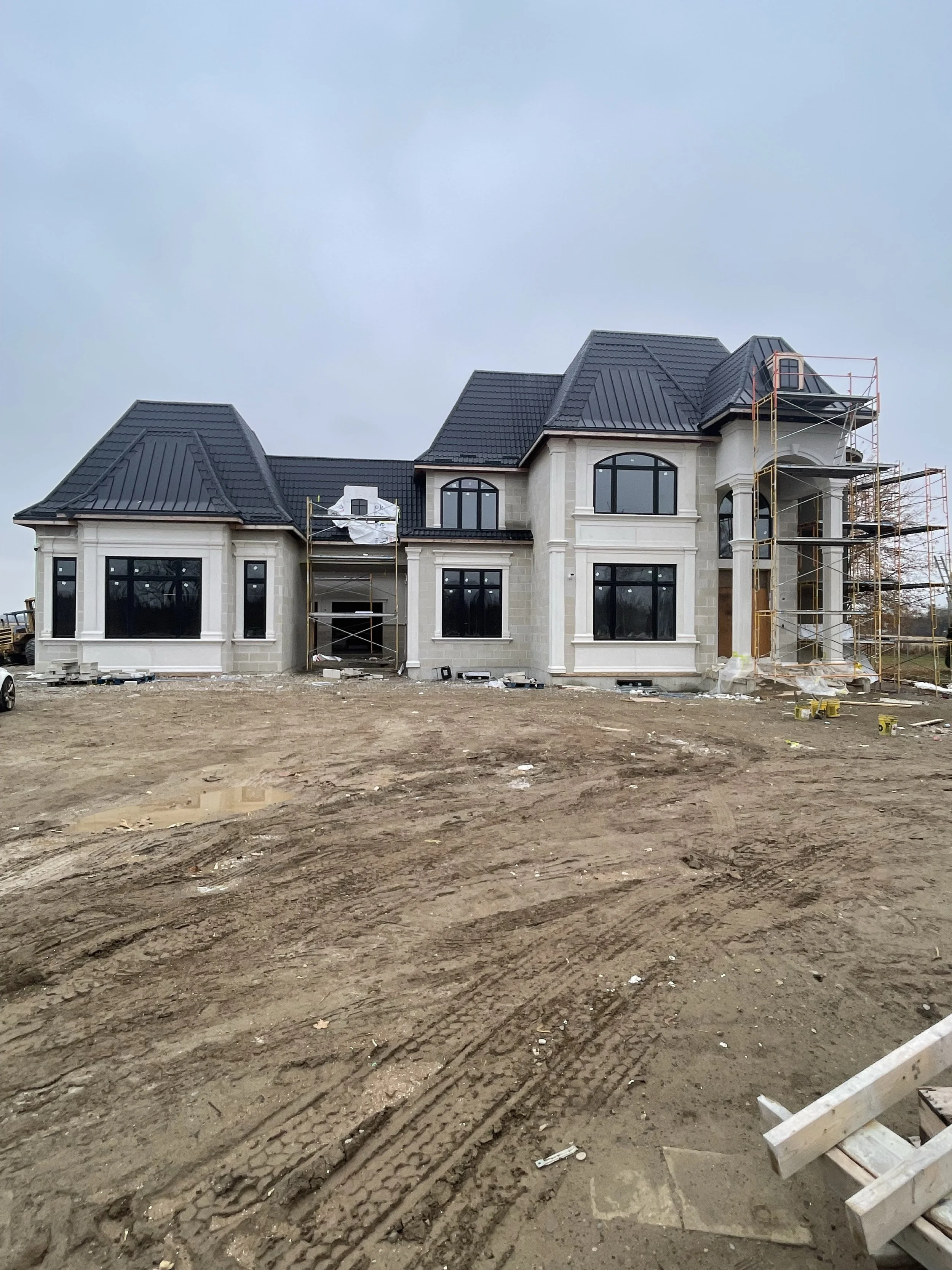 A large, elegant house under construction with scaffolding on the sides, black roof, and multiple windows in a spacious yard.