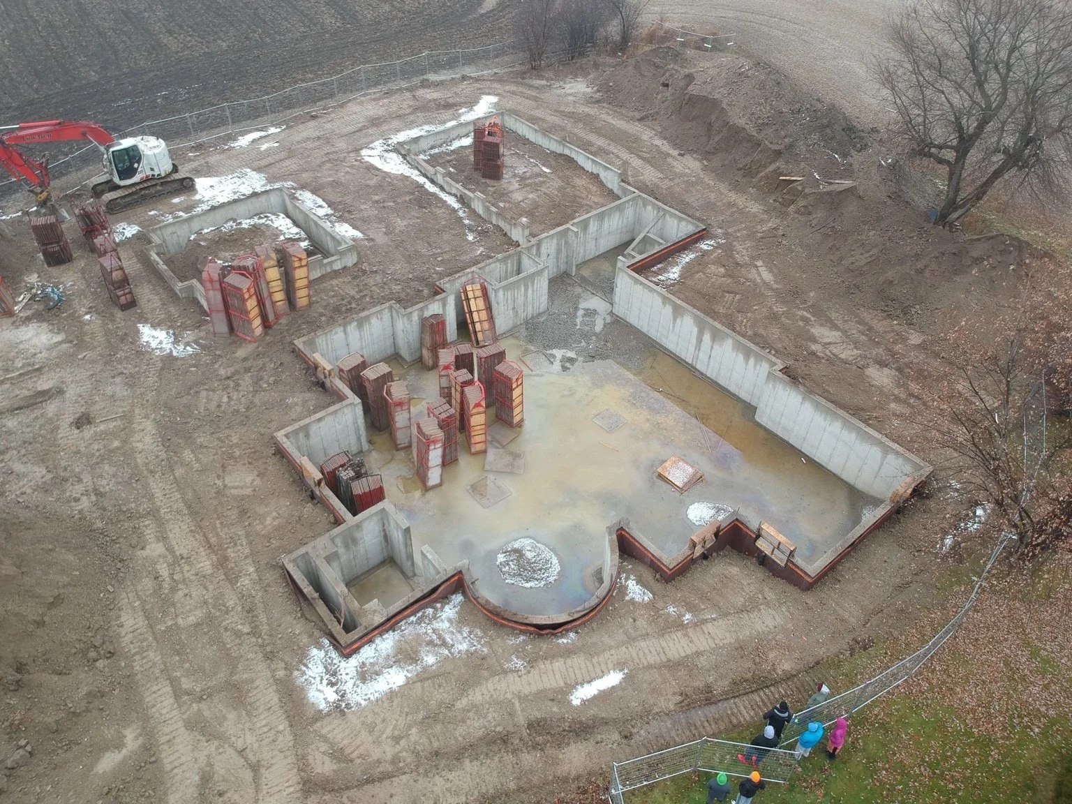 A construction site with concrete foundation walls and metal reinforcements in a muddy area, with a small group of workers or visitors wearing helmets near the fence.
