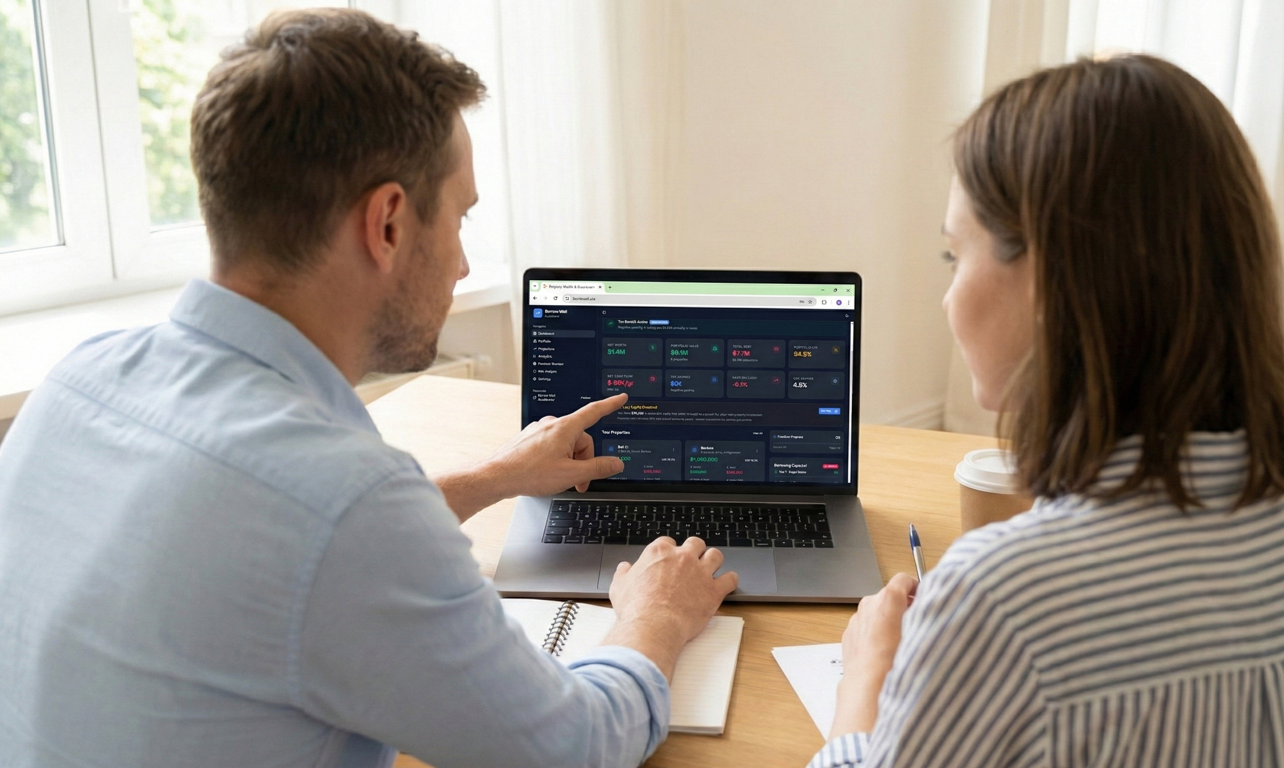 A man and woman sitting at a wooden table, looking at a laptop screen with financial dashboard, with a notebook, pen, and coffee cup on the table.