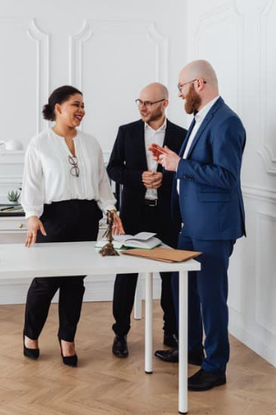 Three professionals engaged in a discussion in a modern office with white walls, a desk with an open notebook, and a small decorative plant.
