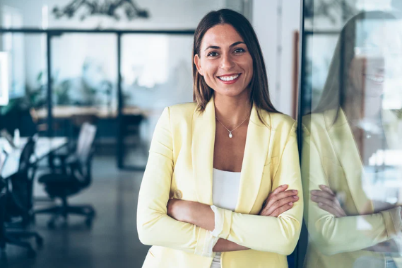 A smiling woman with dark hair wearing a yellow blazer and white top standing with arms crossed in a modern office.