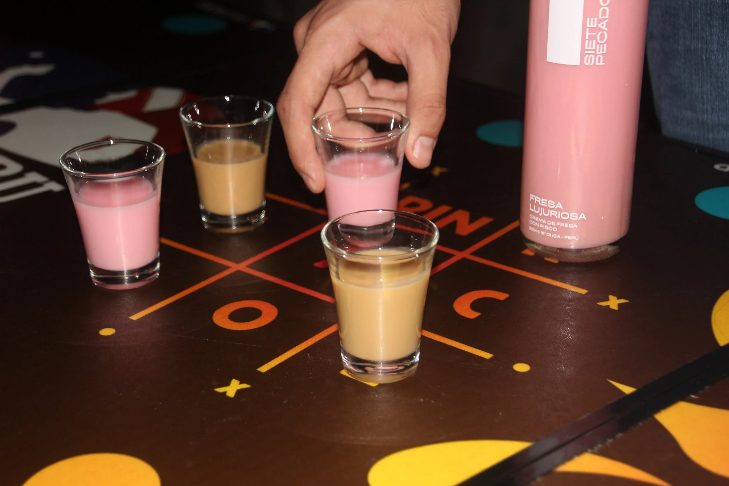 Shots de crema de pisco 7 Pecados en tonos rosado y beige servidos sobre mesa de juego, ambiente nocturno y experiencia divertida para compartir. Fresa Lujuriosa y Pereza de Café.