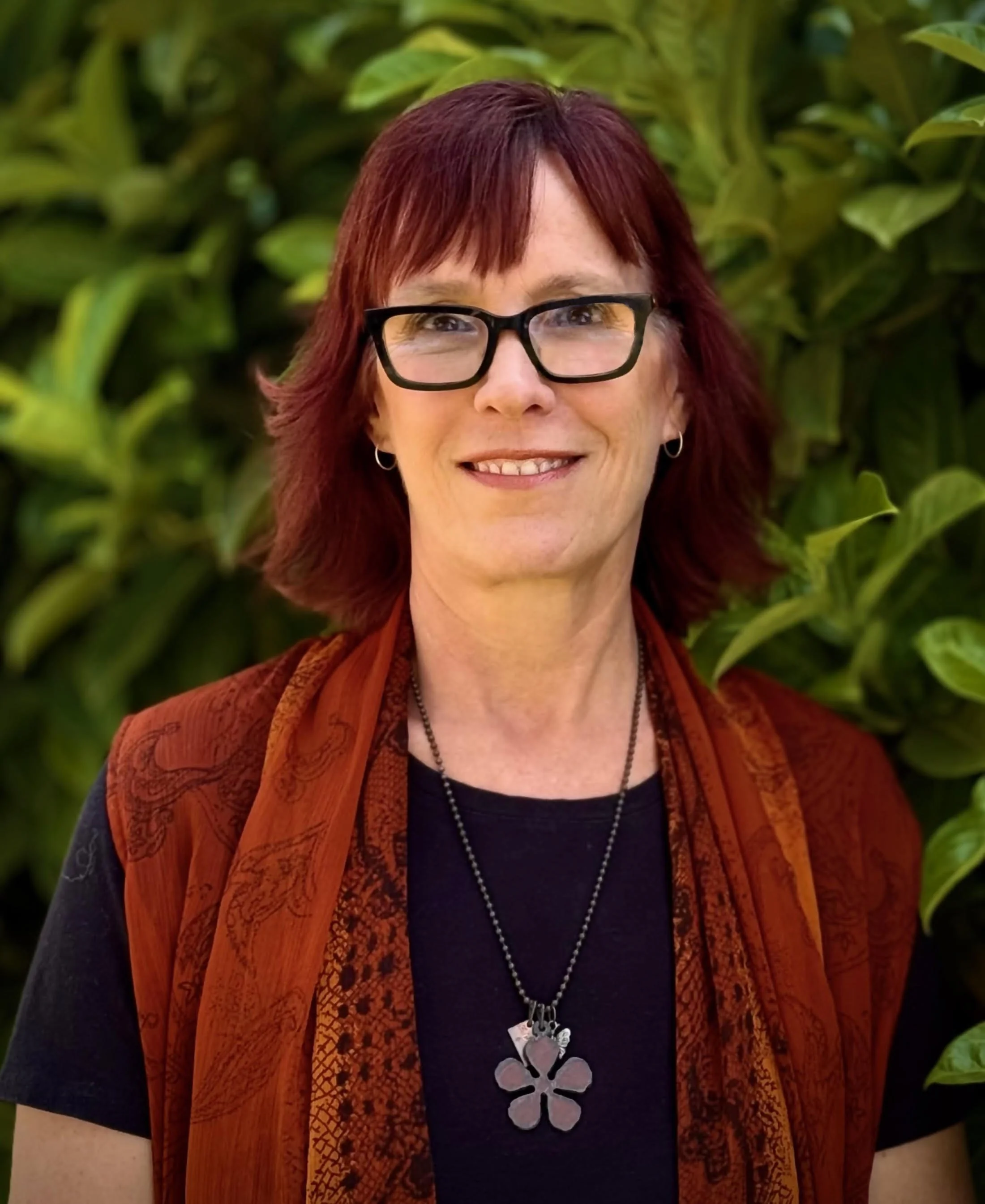 Smiling woman with red hair and glasses standing outdoors in front of green leafy background.