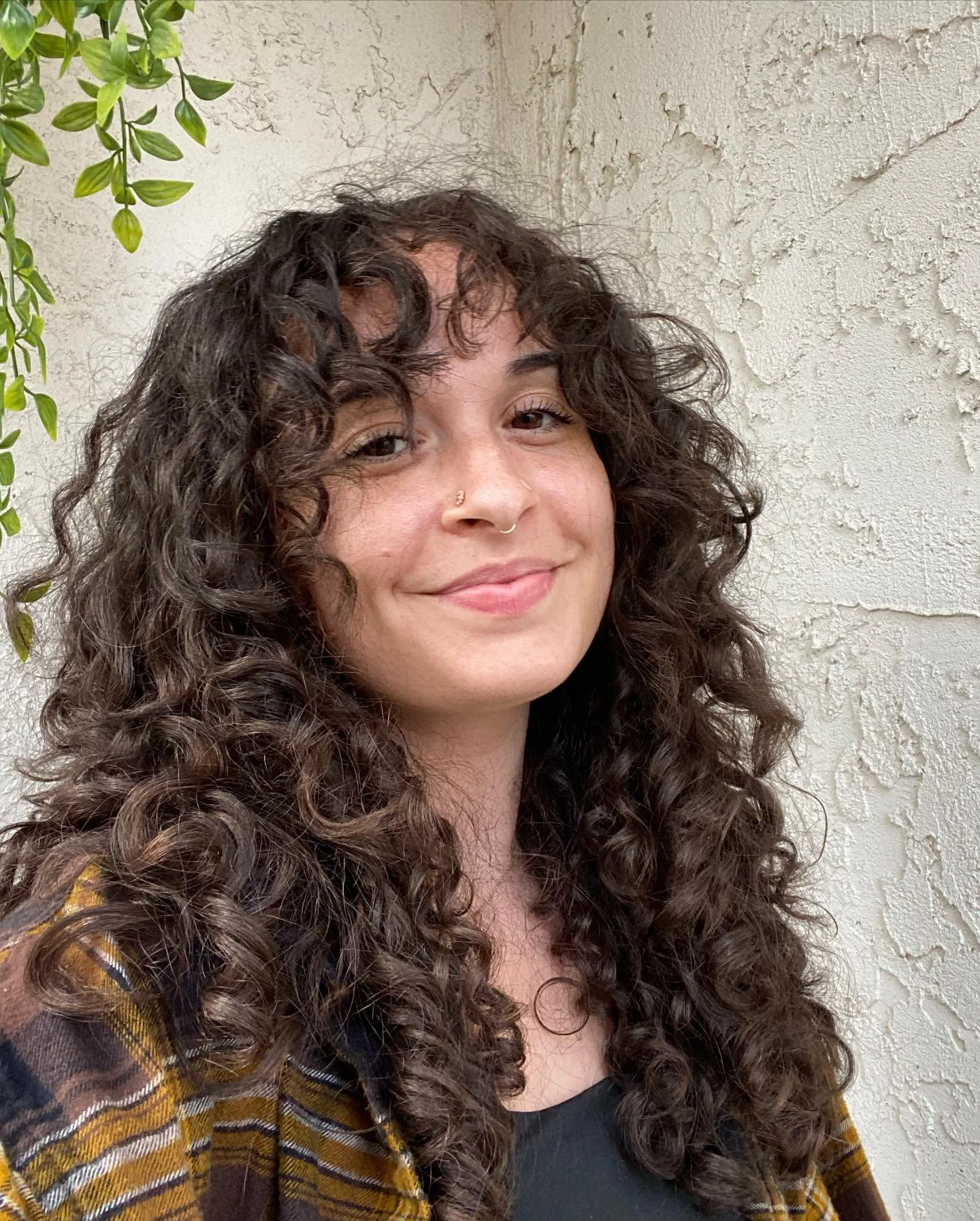 A young woman with curly dark brown hair, light skin, light makeup, and a nose piercing, smiling slightly while facing the camera outdoors near a textured white wall with some green foliage in the background.