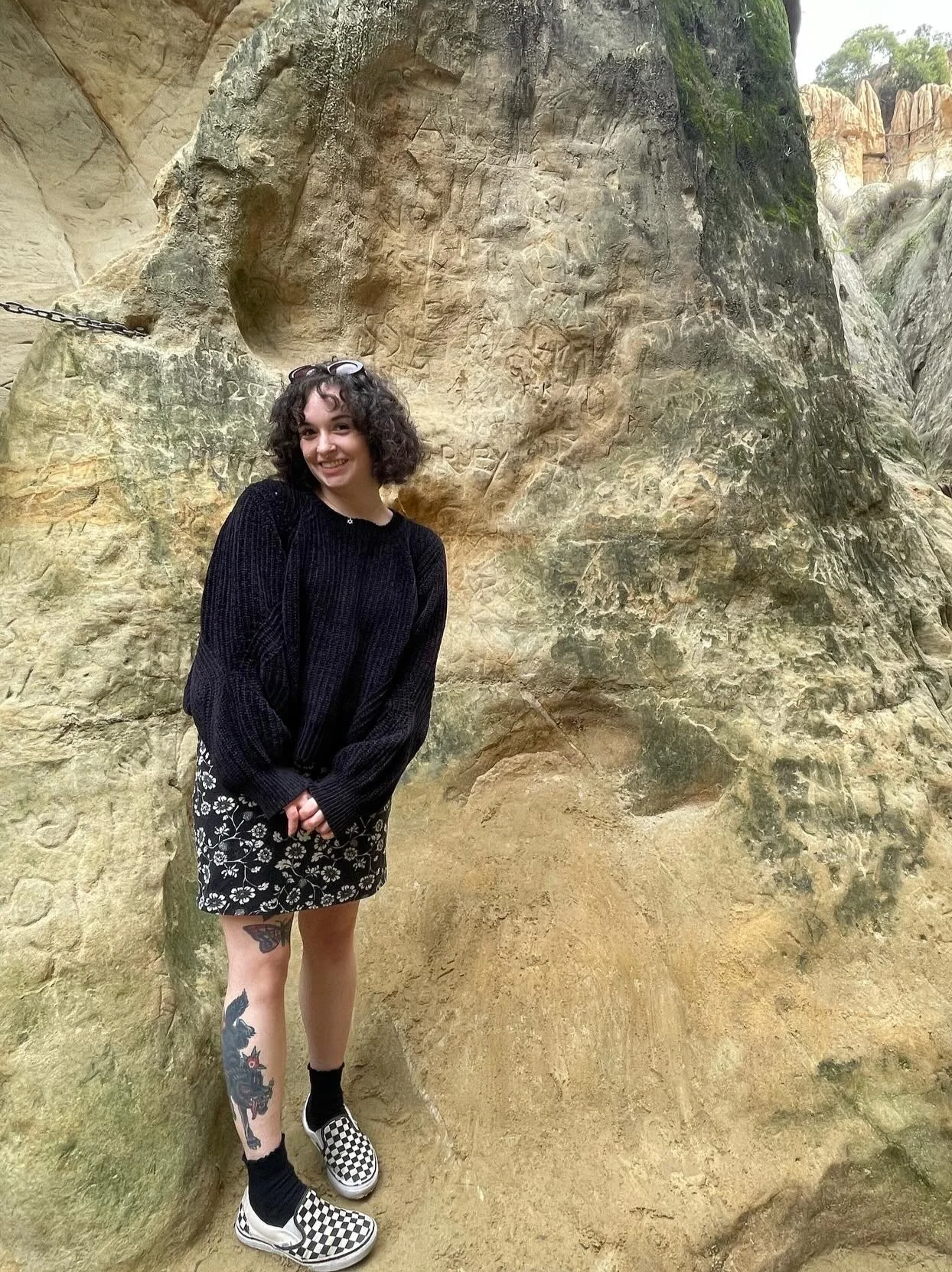 Young woman with curly hair, black sweater, floral skirt, tattoos on her left leg, and checkered Vans shoes standing in front of a large, textured rock formation with carvings.