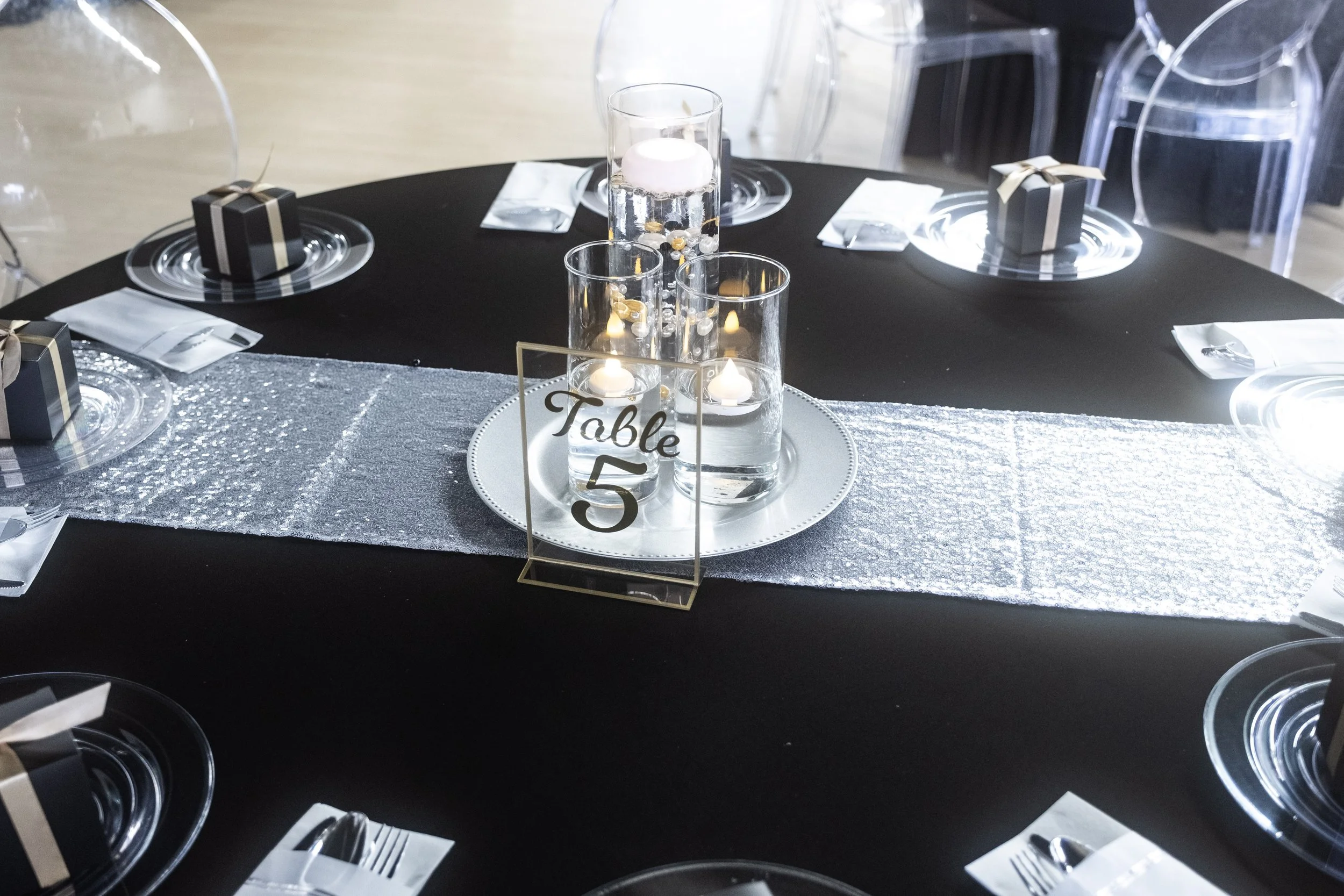 Round black banquet table with white and silver decorations, centerpieces with three candles and a table number '5', set with clear plates, napkins, forks, and small black gift boxes with gold ribbons, surrounded by transparent chairs.
