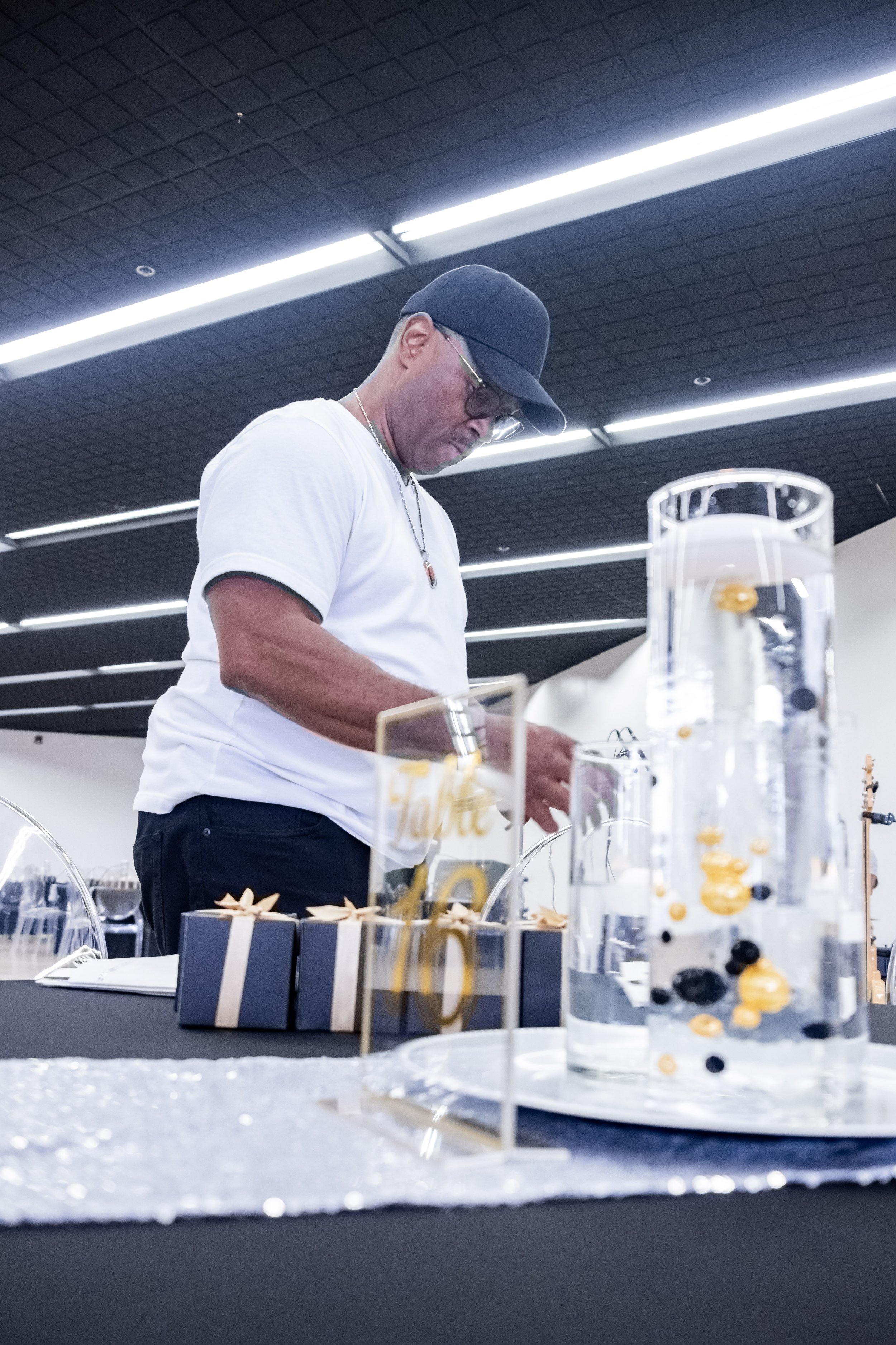 A man wearing a white t-shirt, black cap, glasses, and a necklace is standing at a table with decorative items and glass containers filled with water and gold and black objects. The background shows a black ceiling with bright, linear lighting.