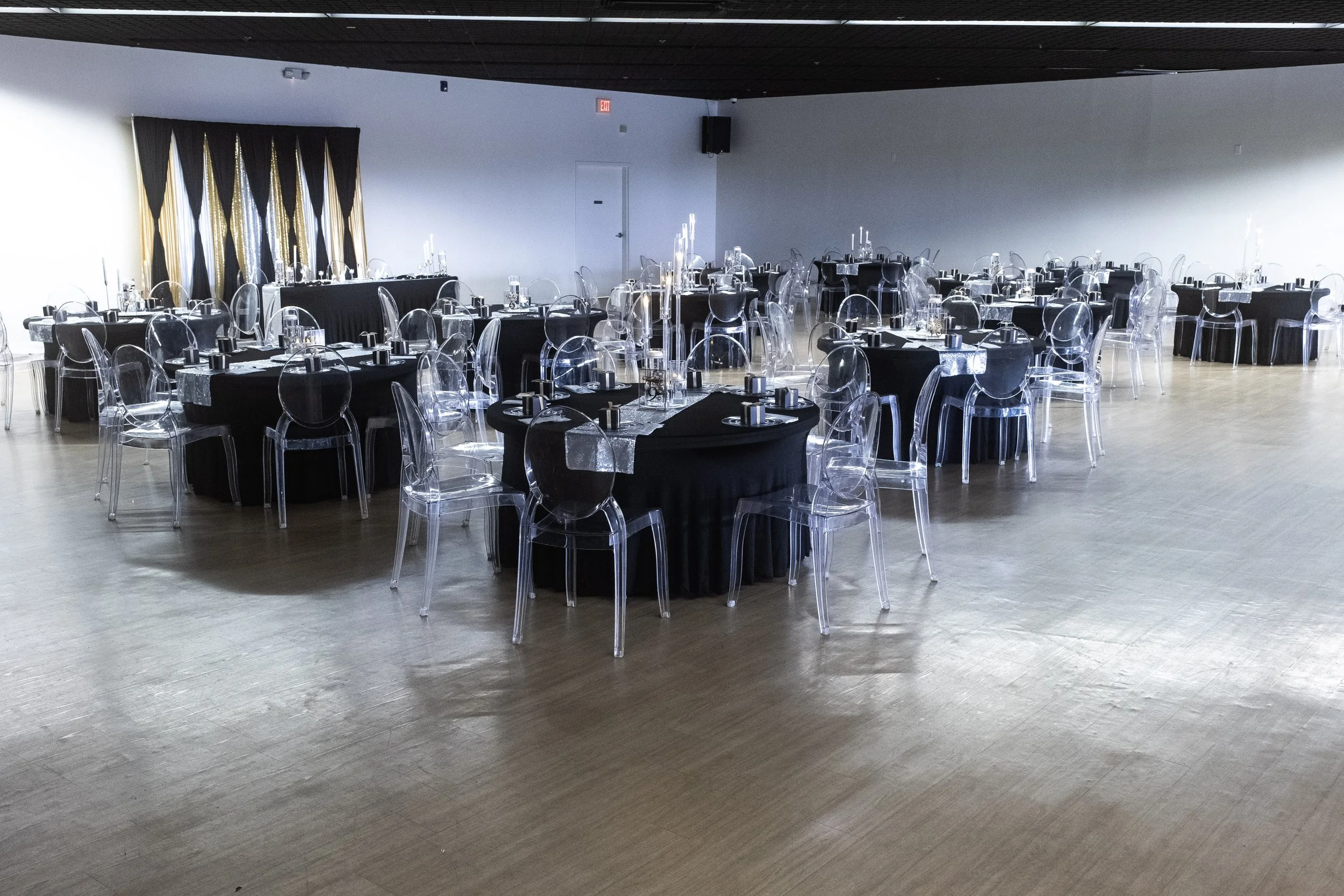 Banquet hall with round tables covered in black tablecloths, transparent ghost chairs, and elegant table settings with glassware and candles, prepared for an event.