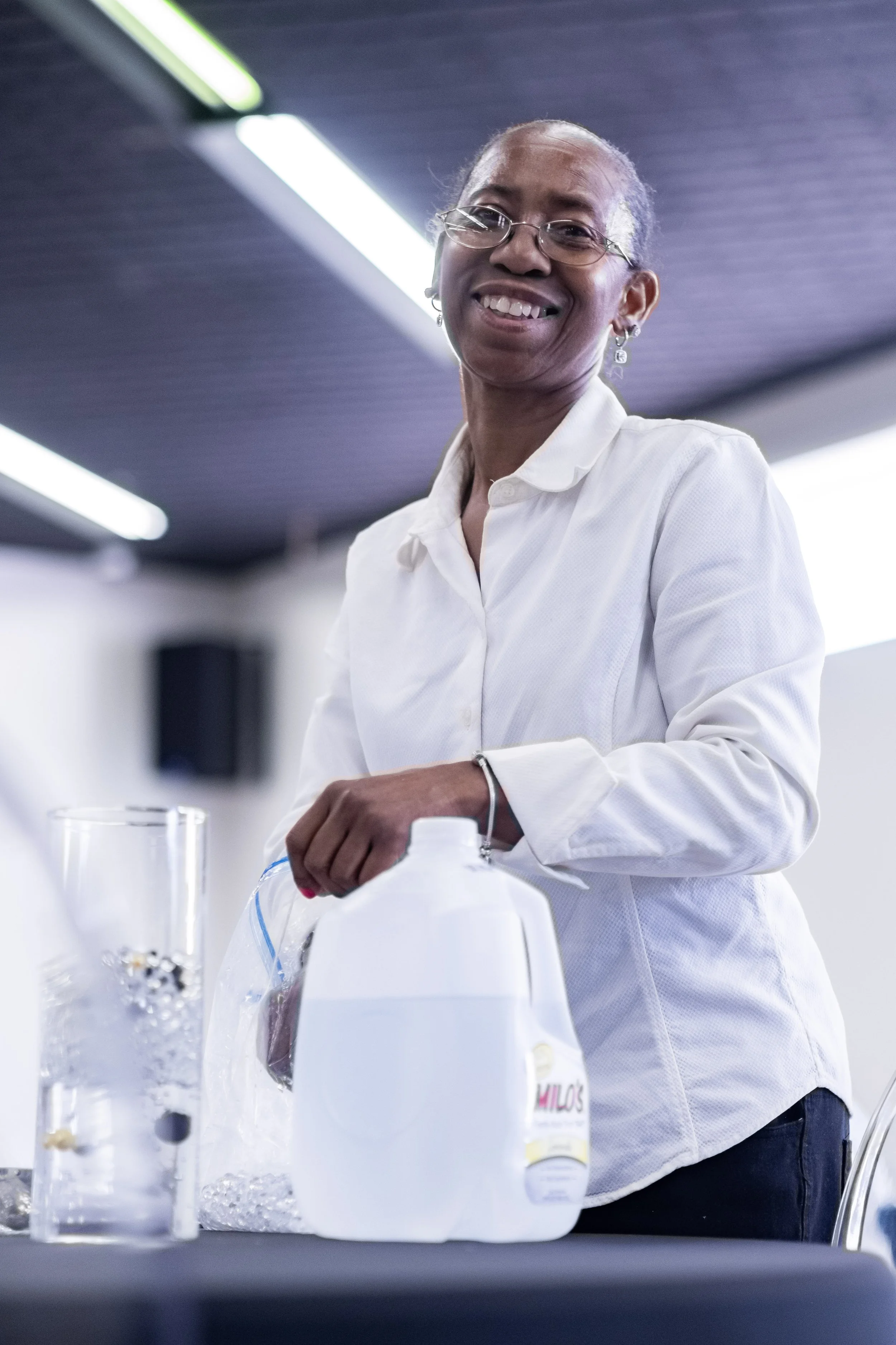 A smiling woman with glasses and earrings standing at a table with a gallon jug and other laboratory equipment in a bright, modern room.