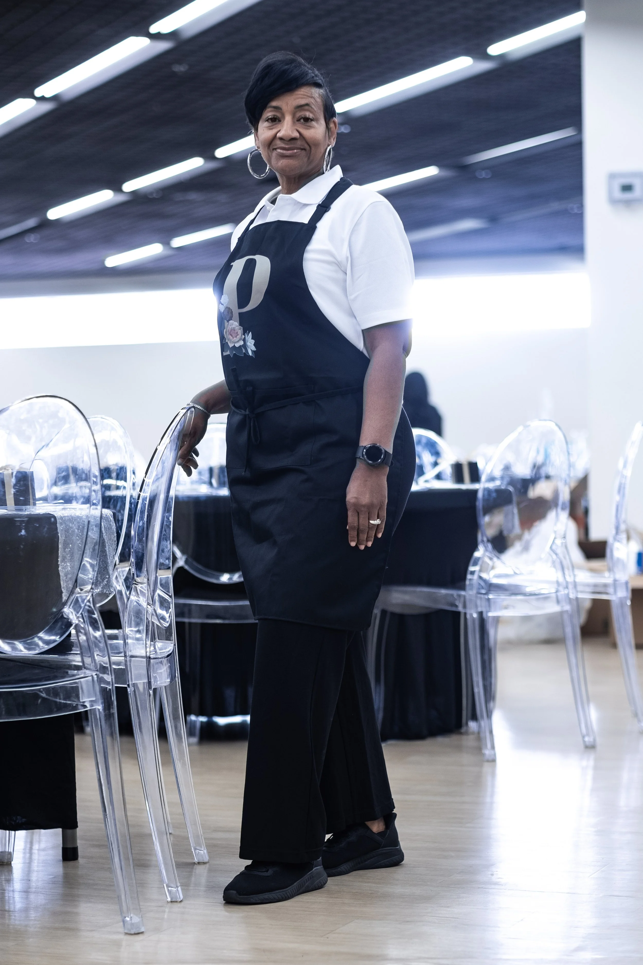 A woman stands in a modern dining area, wearing a white shirt and black apron, with a confident expression.