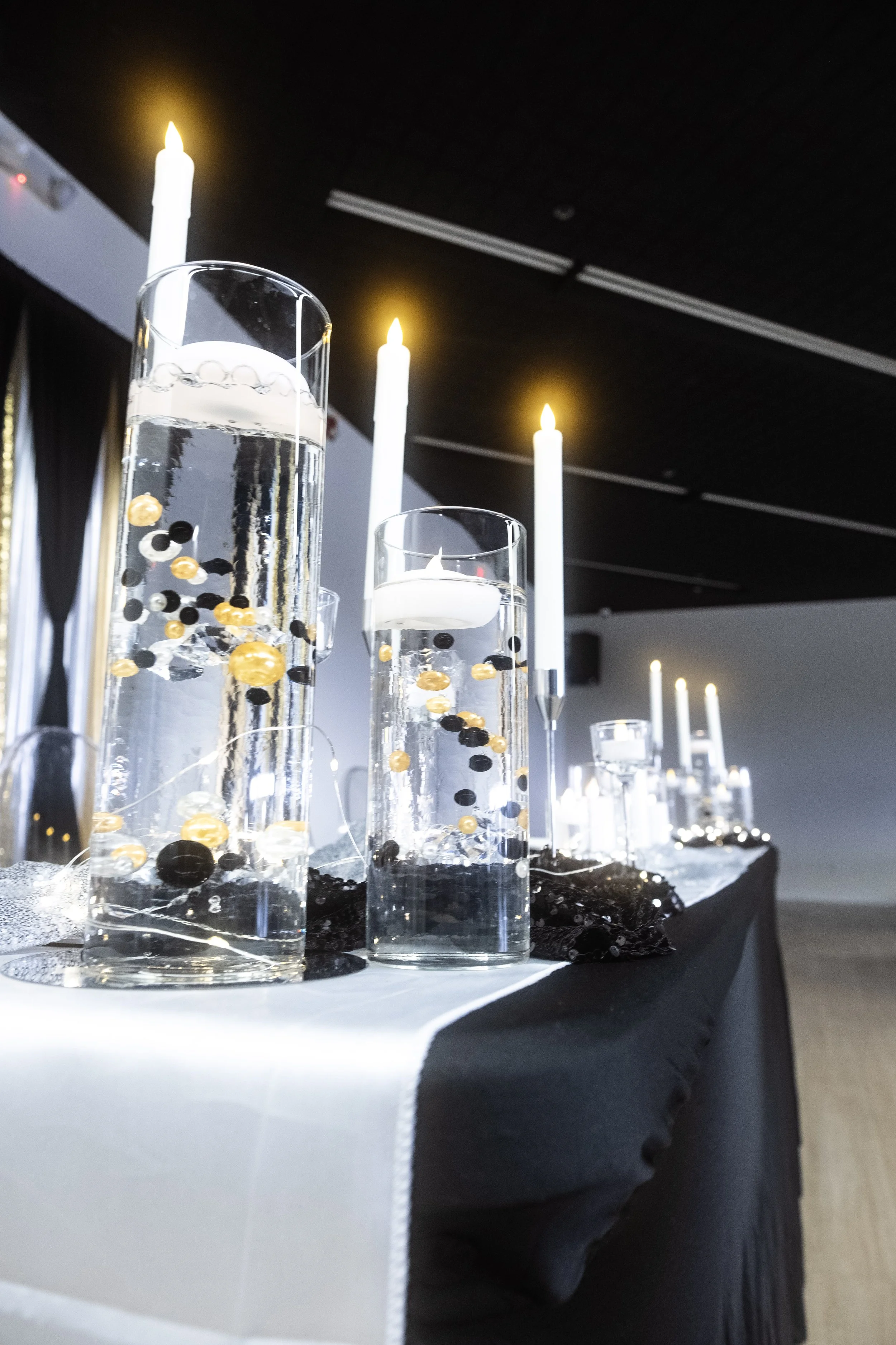 Table decorated with tall glass vases filled with water and black and gold beads, lit white candles, black and silver fabric, and string lights, set in a dimly lit room with black ceiling and dark curtains.