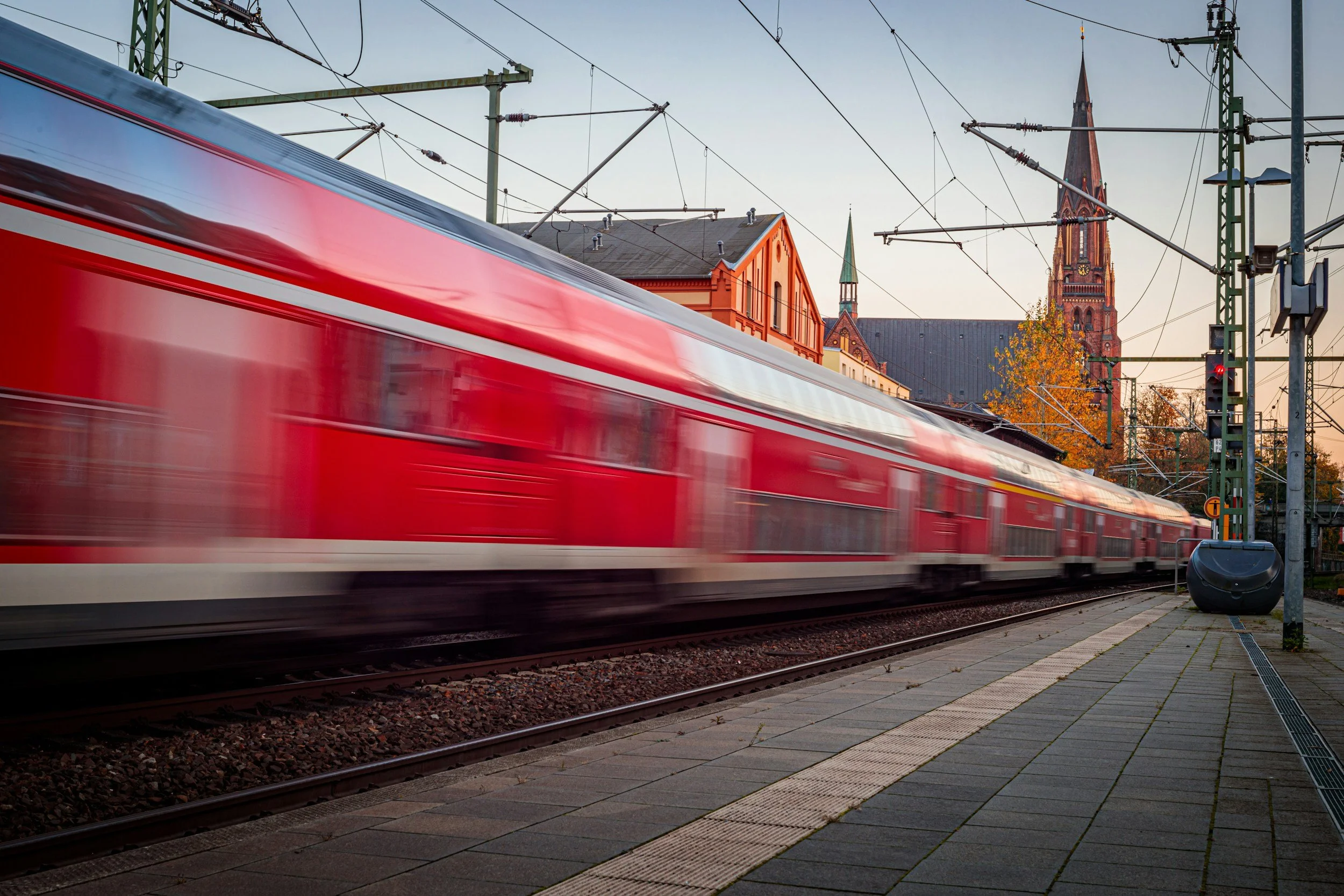 A red train moving quickly along railway tracks with a church and other buildings in the background at sunset.