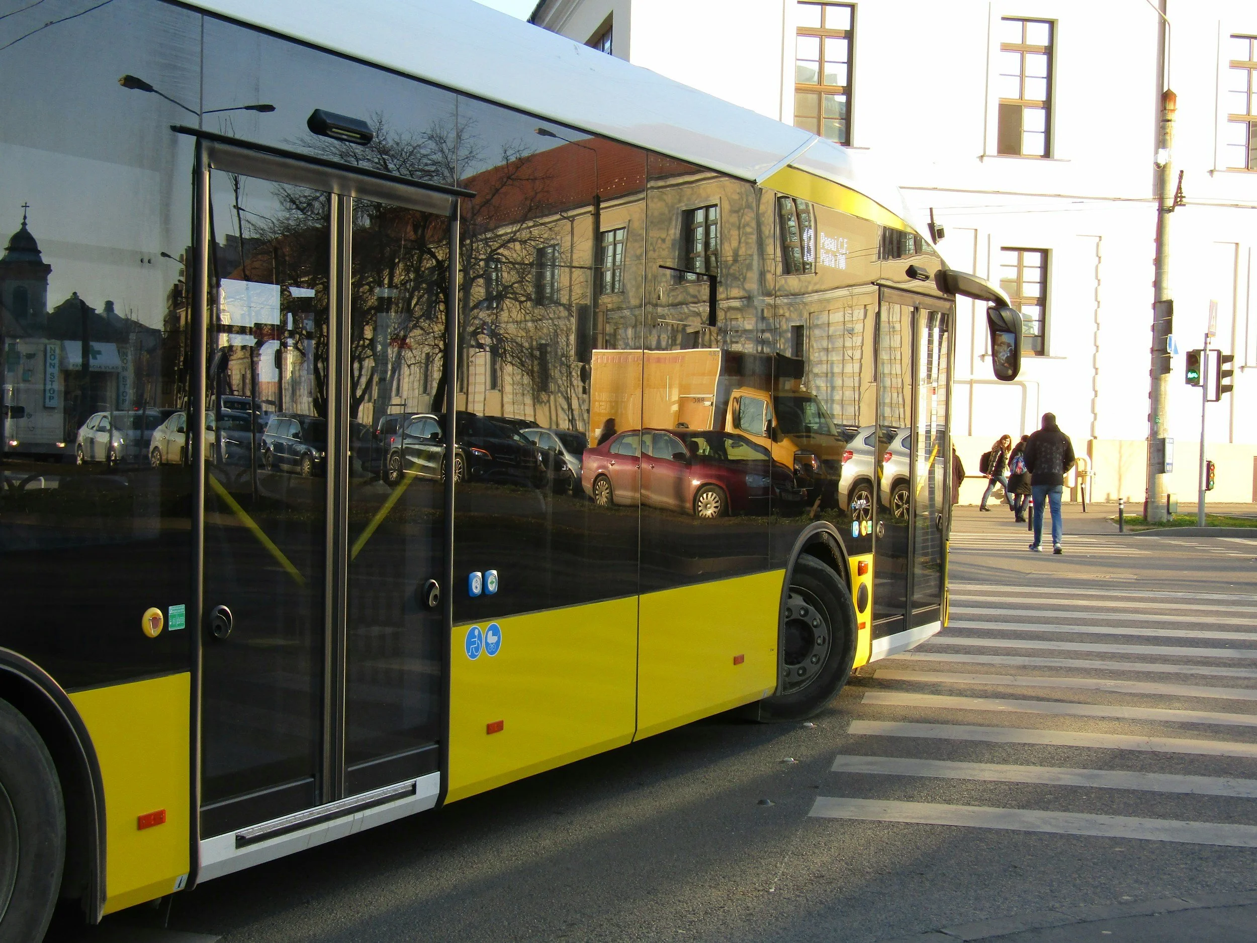 A yellow and black city bus stopped at a crosswalk, with pedestrians walking nearby and cars reflected on its shiny, tinted windows.