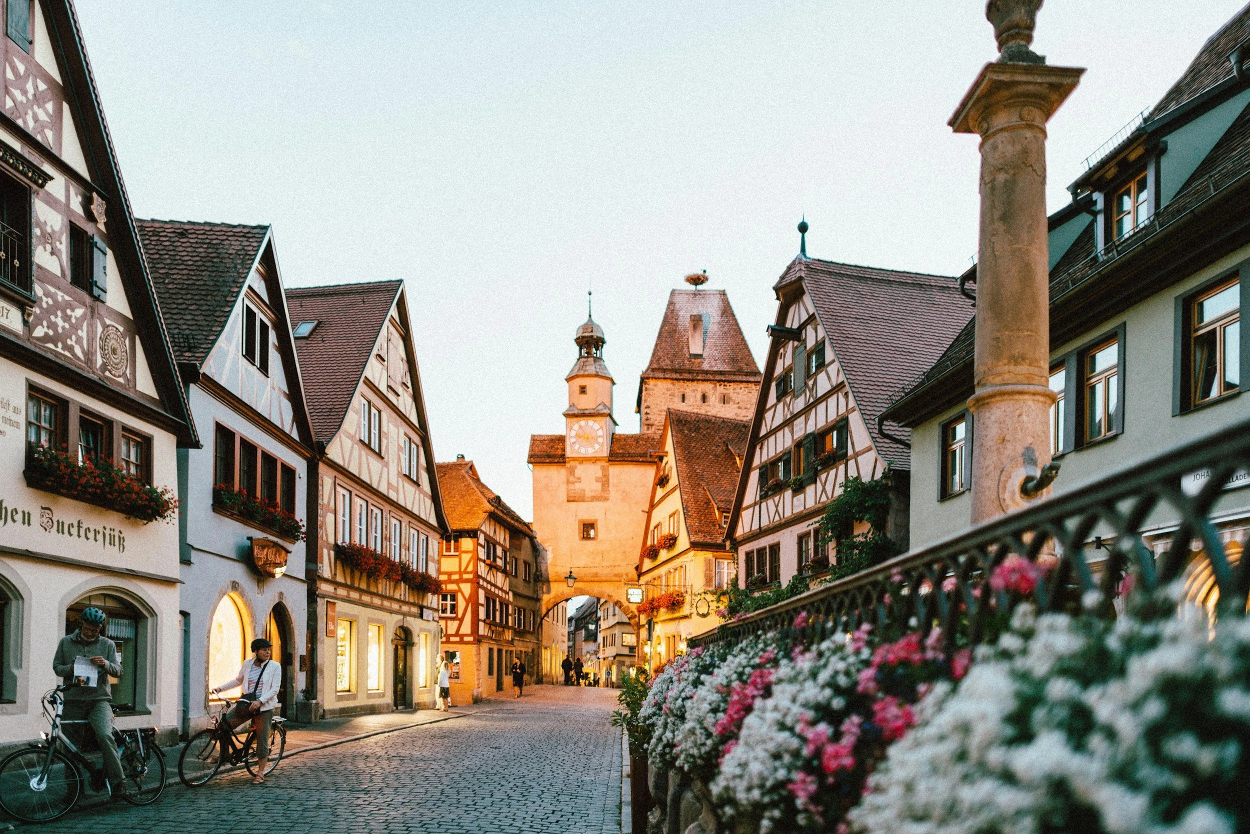 A European street scene at sunset with colorful historic buildings, a clock tower, pedestrians, and flowers in the foreground.