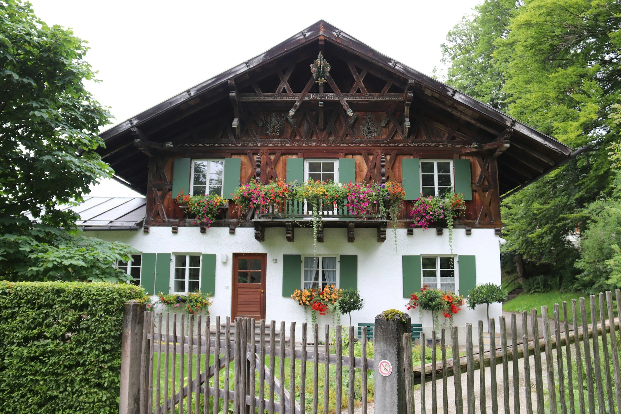 Traditional two-story house with a white lower level, wooden upper level with green shutters, flower boxes on windows, and a wooden fence in the front yard surrounded by greenery.