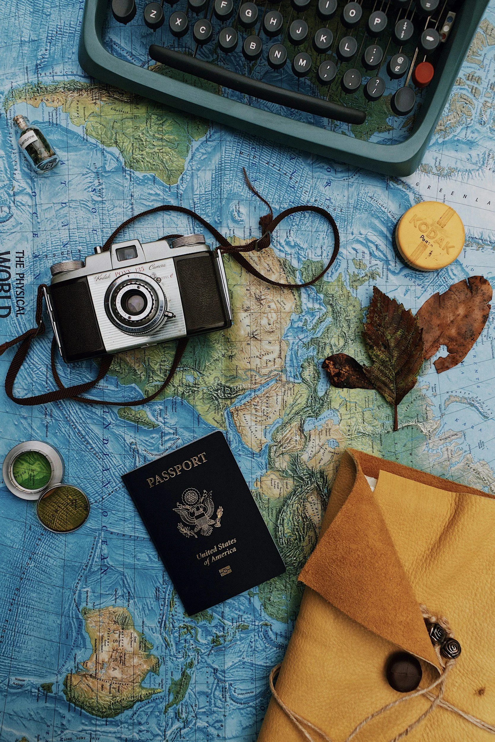 Travel essentials on a world map including a vintage camera, a U.S. passport, a Kodak film container, a small bottle of perfume, a magnifying glass, a compass, leaves, a yellow envelope, and a typewriter.