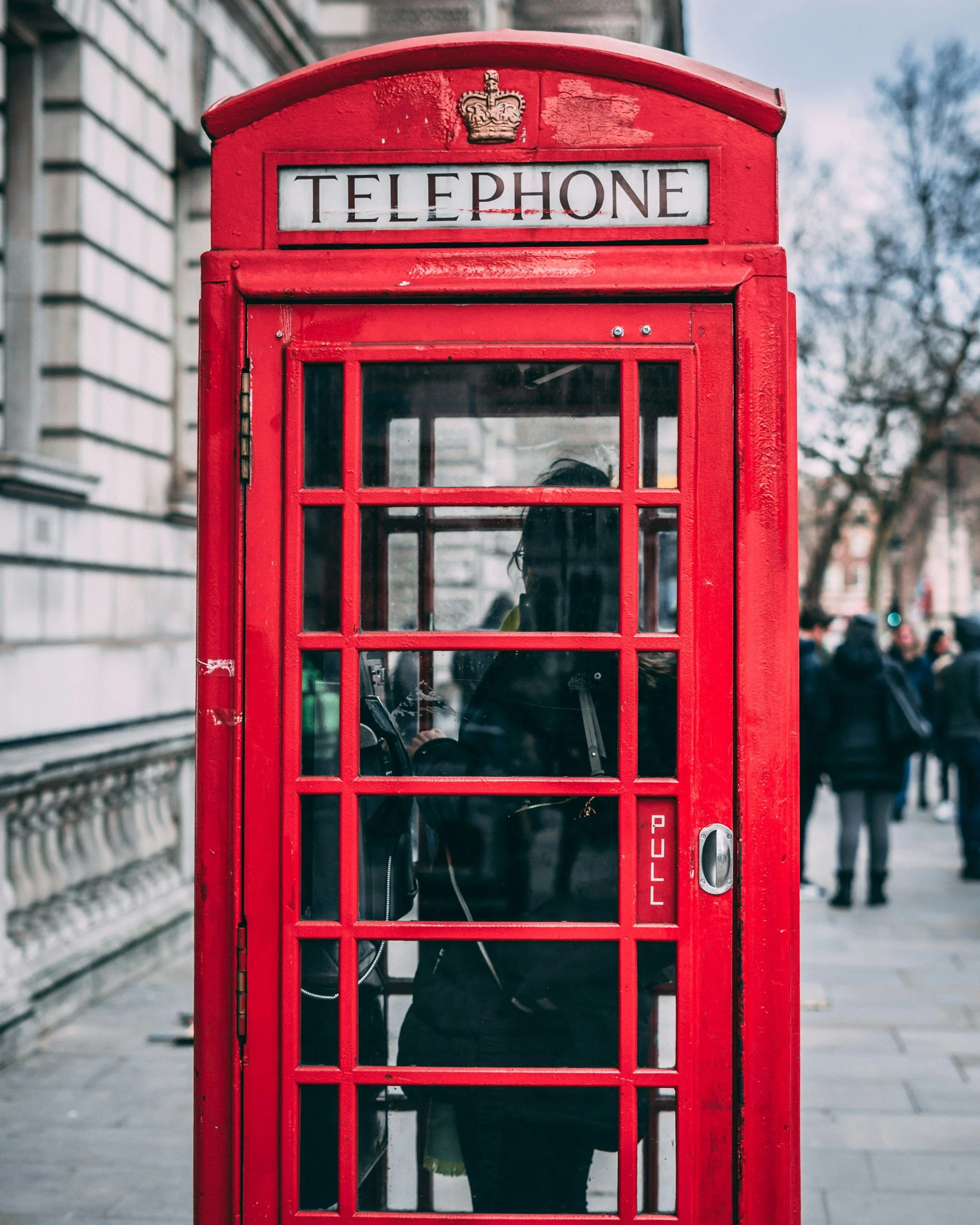 Red British telephone booth on a city sidewalk with people walking in the background.