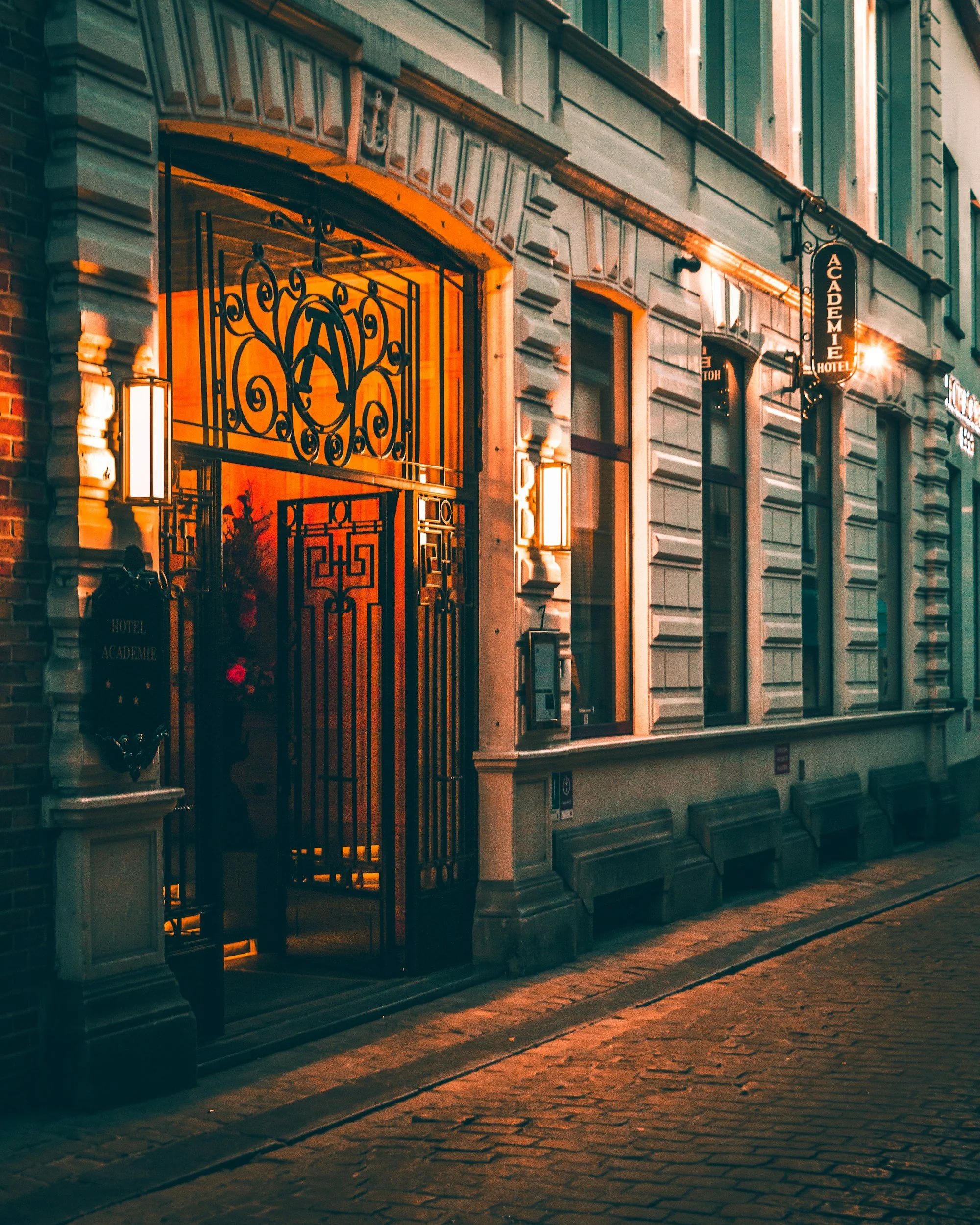 Night view of the entrance to Hotel Académie with illuminated sign, ornate black gate, warm lighting, and cobblestone street outside.