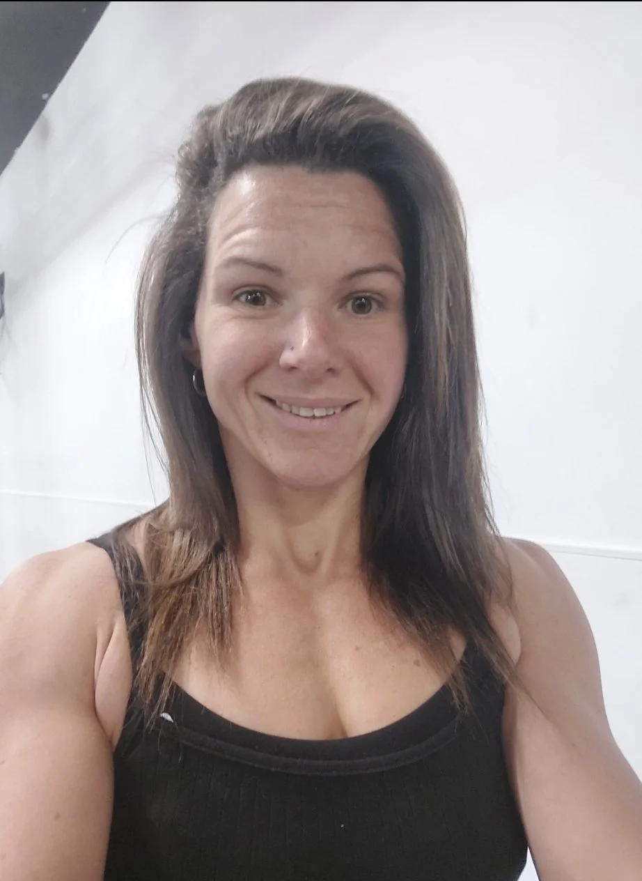 A woman with shoulder-length brown hair, wearing a black tank top, smiling for a selfie in front of a white background.