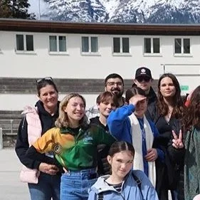 Group of seven friends posing outdoors in front of a building, some making peace signs, with a hillside and trees in the background.