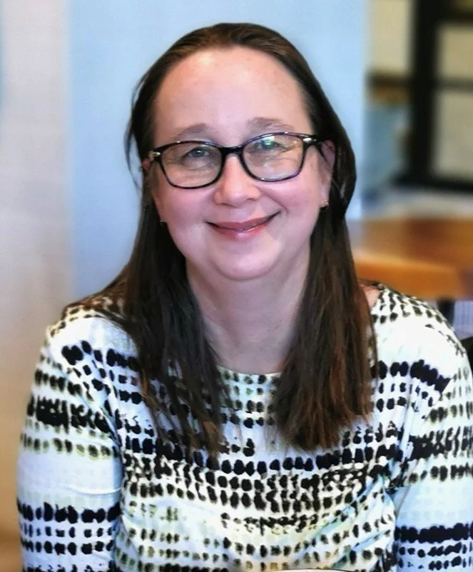 A woman with long dark hair, wearing glasses, smiling, in a patterned black and white top, sitting indoors.