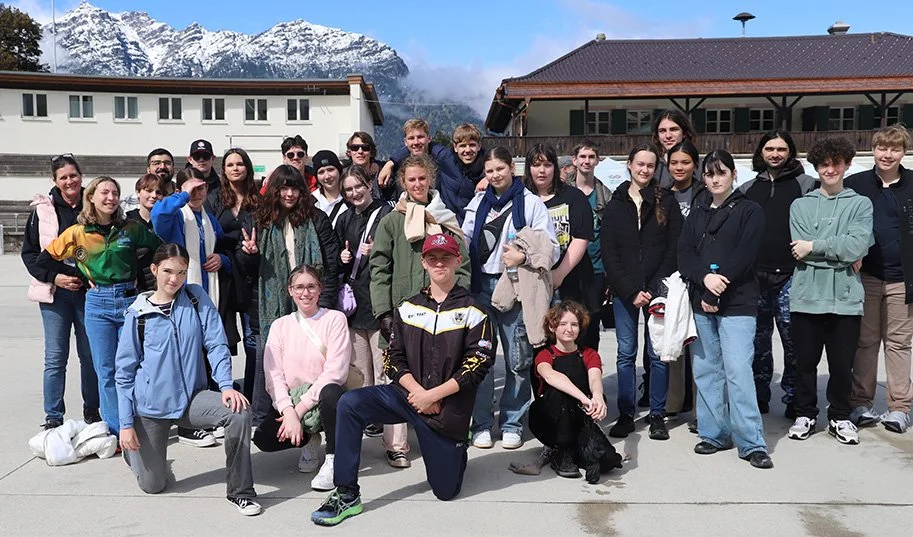 Group of young people standing outdoors in front of a building with mountains in the background, some people are smiling and making peace signs.