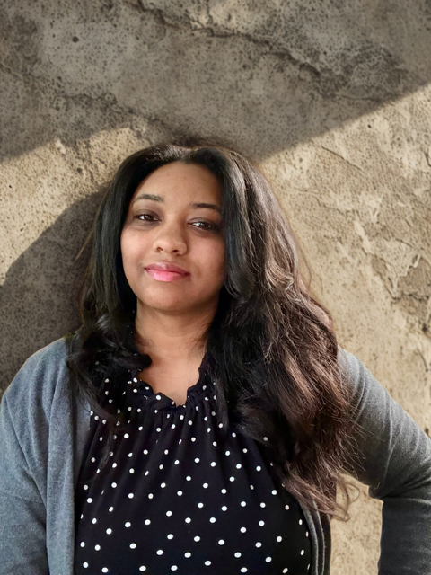 A woman with dark hair, wearing a polka dot blouse and a gray cardigan, standing against a textured wall, looking at the camera with a slight smile.