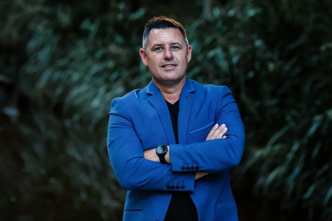 A man wearing a blue blazer with arms crossed, standing outdoors with blurred green foliage in the background.