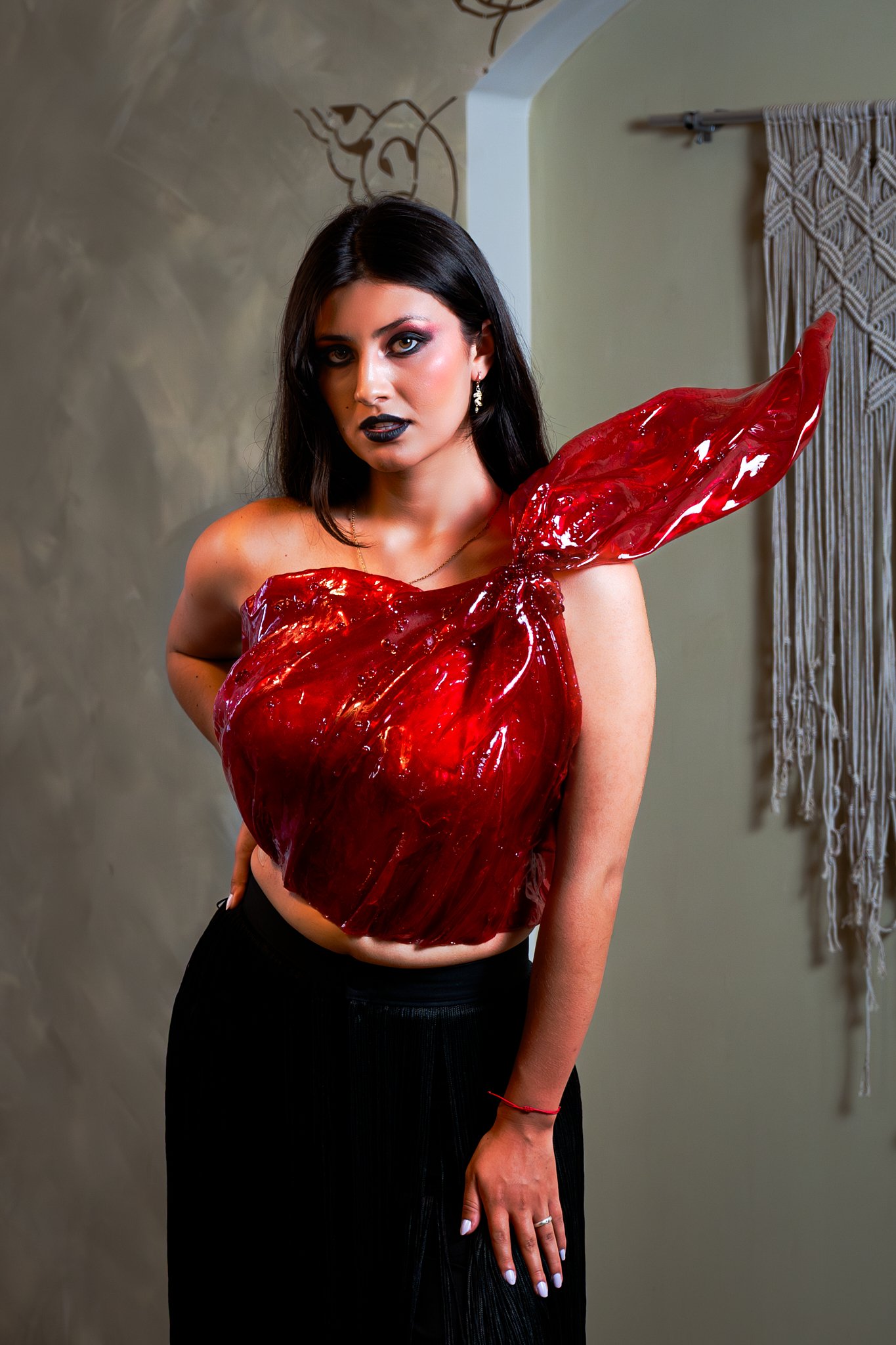 Mujer con maquillaje oscuro y vestido de estructura de color rojo brillante y transparente, con un diseño inspirado en una flor o capullo, en un fondo neutral con cortina de macramé.