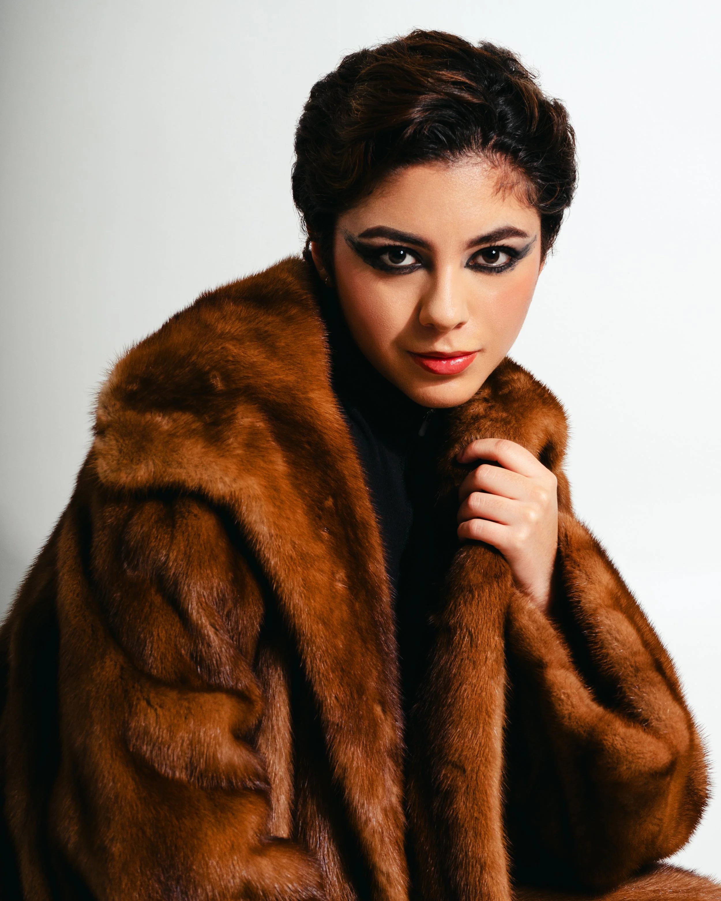 Mujer con maquillaje oscuro y abrigo de piel marrón, mirando a la cámara con expresión seductora.