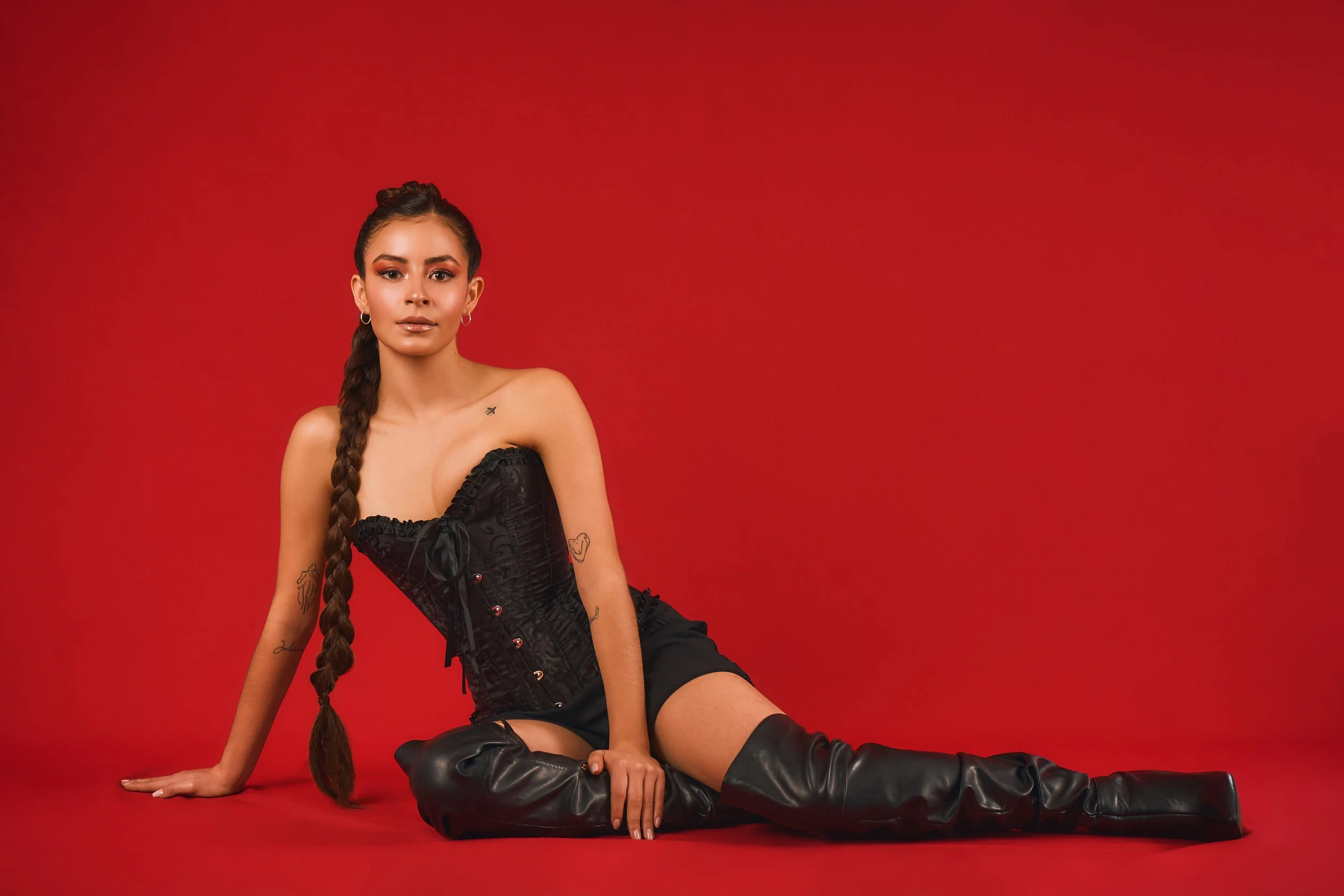 Joven mujer en vestido negro y botas largas sentada en el suelo rojo, con fondo rojo, mirada al frente, cabello largo trenzado.