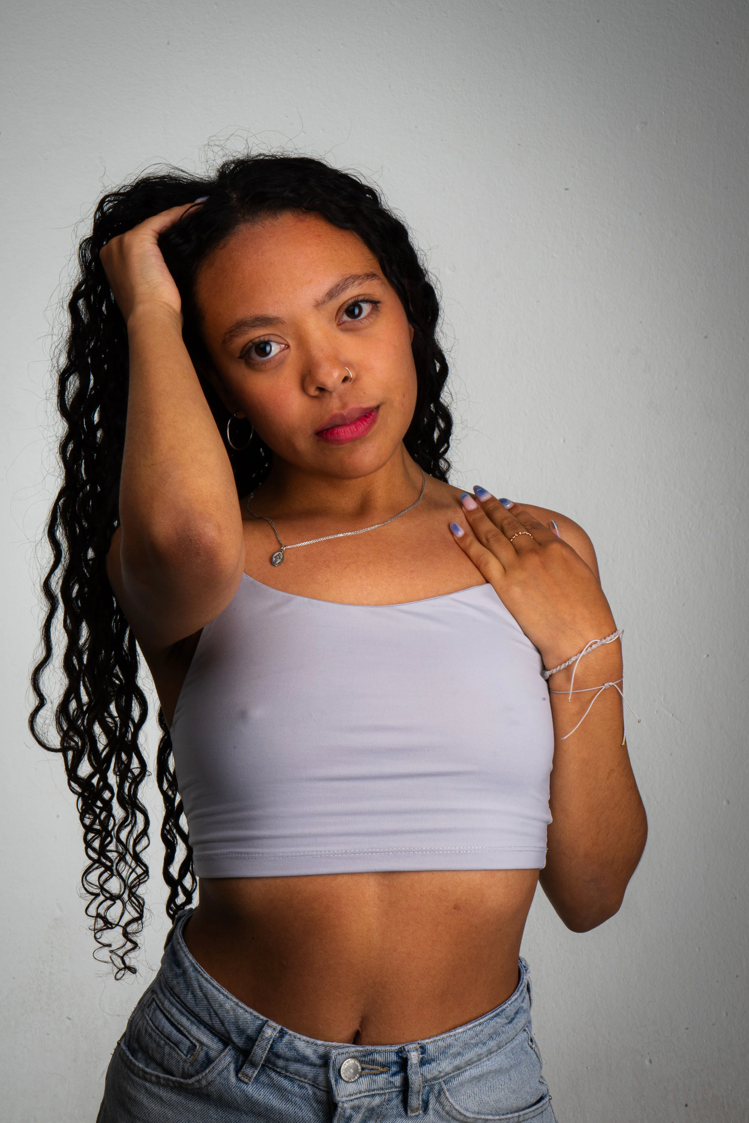 Joven mujer con cabello rizado oscuro, sin camiseta blanca, pantalones vaqueros y accesorios plateados, posando frente a una pared gris, tocándose el cabello y la mano en el hombro.