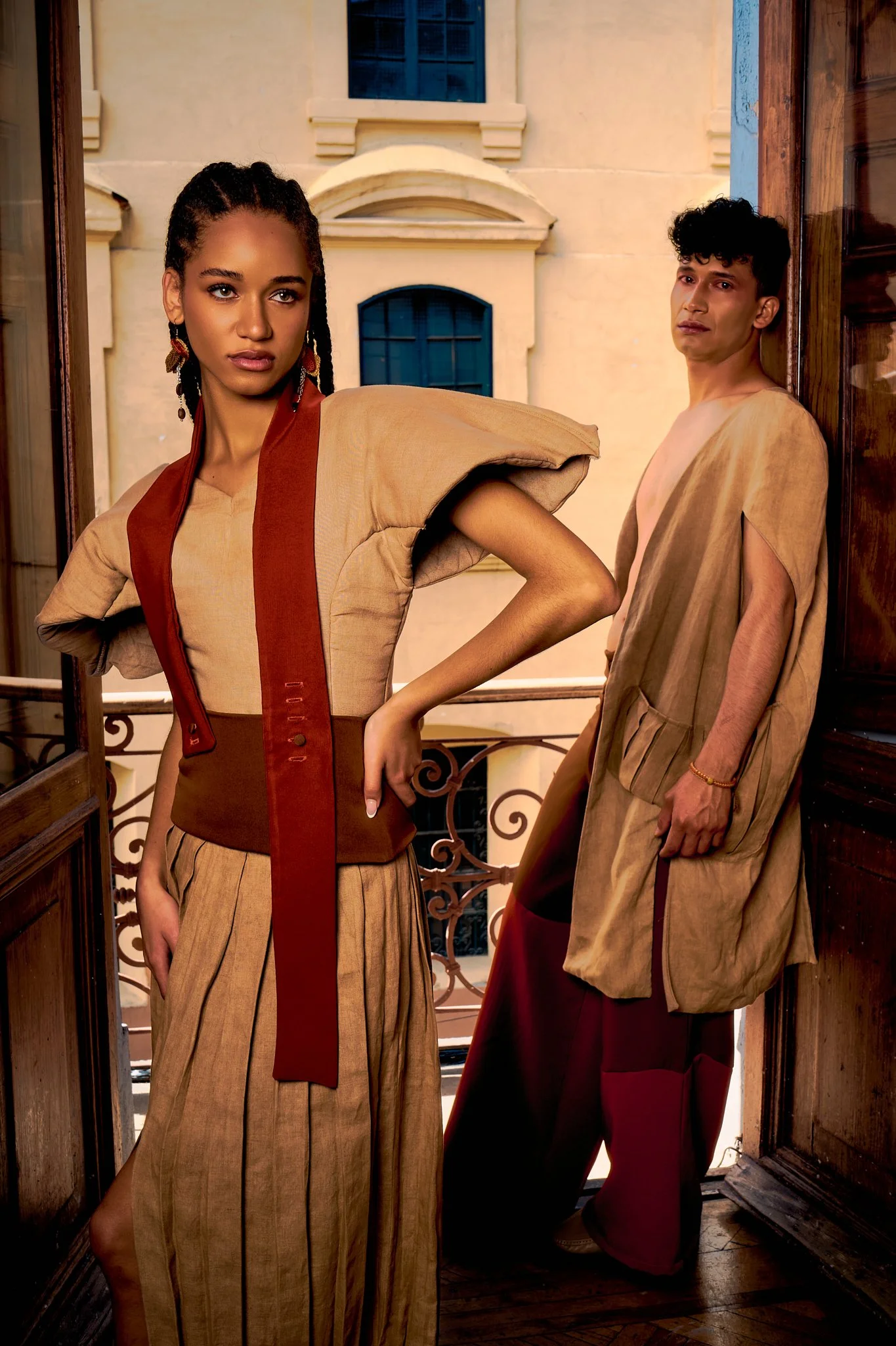 Dos personas con ropa de estilo vintage en un balcón, uno de pie y otro apoyado en la puerta, fondo de edificio con ventanas azules.